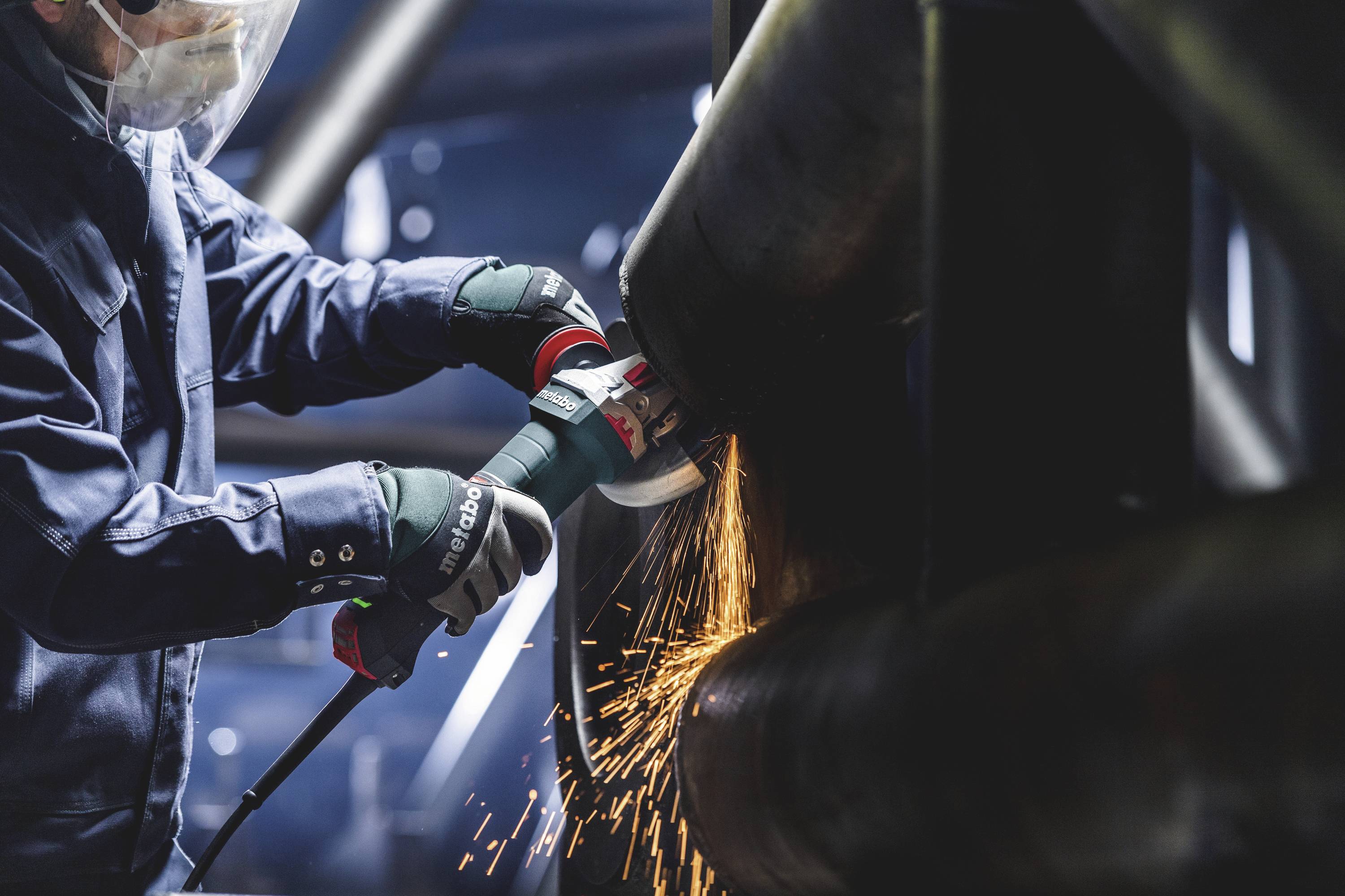 Arbeiter schleift Metallrohr mit Funken. Trägt Schutzkleidung und -visier in industrieller Umgebung. Sicherheit im Vordergrund.