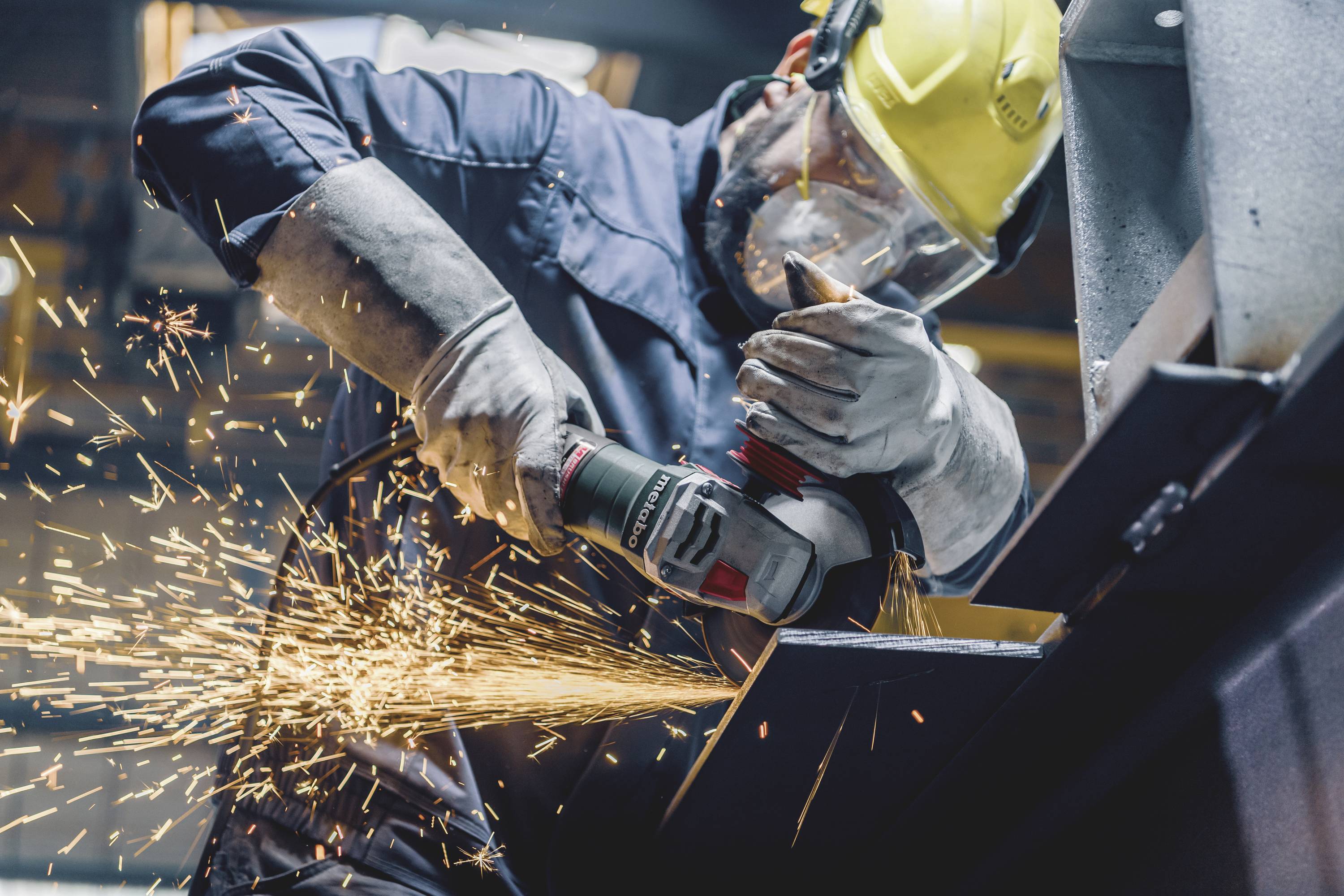 Arbeiter in Schutzkleidung schleift Metall mit einer Schleifmaschine, Funken sprühen.