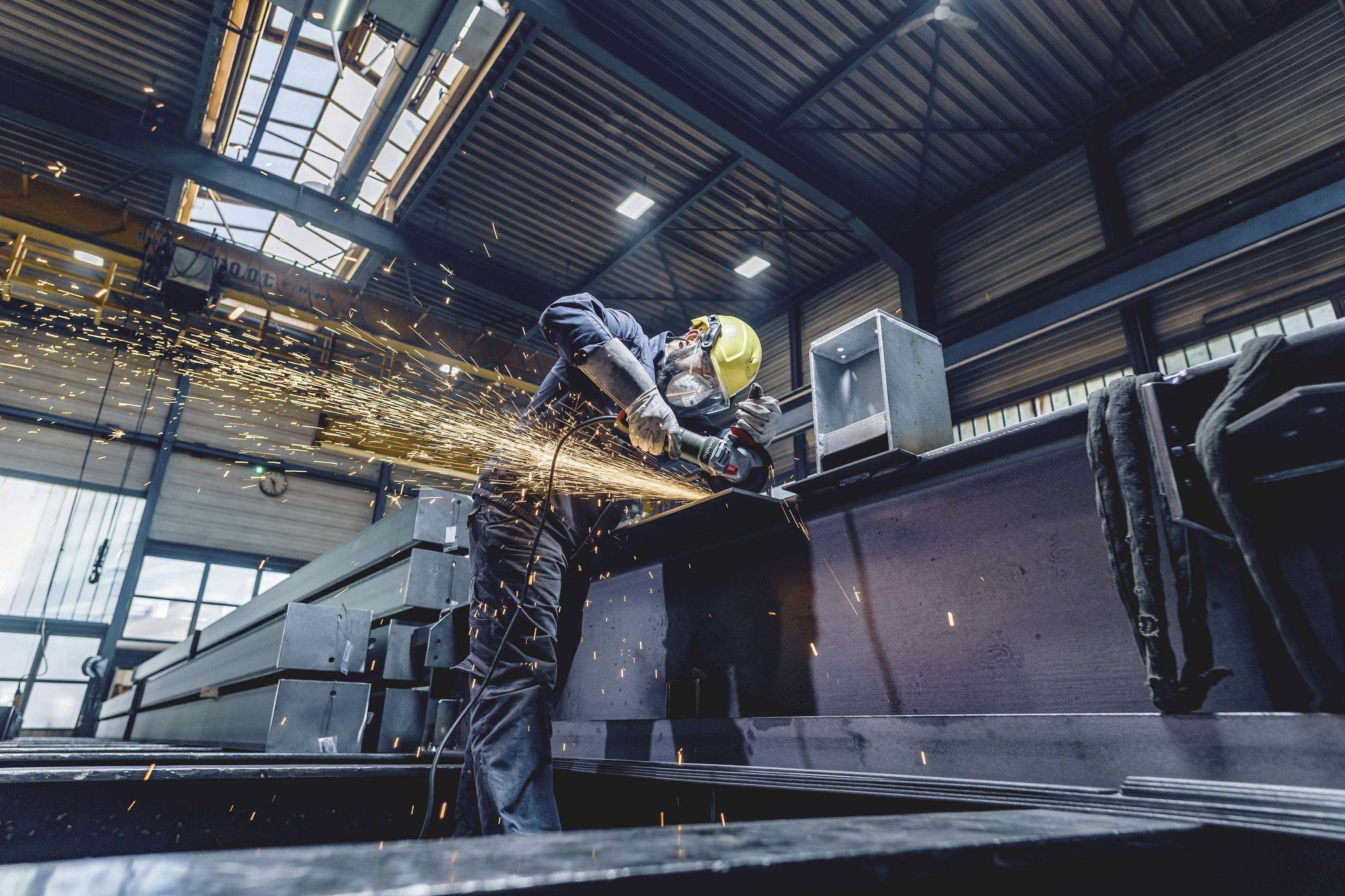 Ein Arbeiter mit Schutzhelm schleift Metall in einer Industriehalle. Funken fliegen, während er ein großes Metallstück bearbeitet.