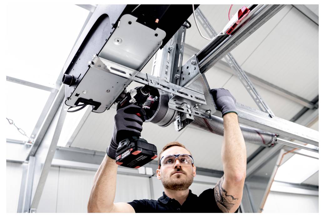 Ein Techniker, der Schutzbrille und Handschuhe trägt, verwendet einen Elektroschrauber, um einen an der Decke montierten industriellen Garagentormechanismus zu installieren oder zu reparieren.