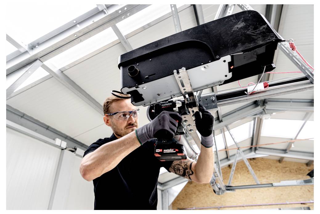 Ein Techniker trägt eine Schutzbrille und Handschuhe und verwendet ein Elektrowerkzeug, um ein an der Decke montiertes Industriegerät in einer Werkstatt zu installieren oder zu reparieren.