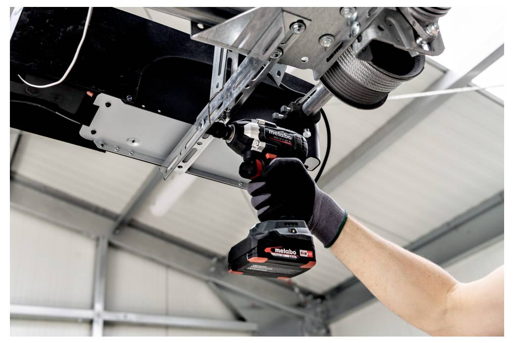 Ein handschuhtragender Techniker benutzt eine kabellose Bohrmaschine, um an einer über Kopf angebrachten Metallmaschine in einer Industriewerkstatt zu arbeiten.