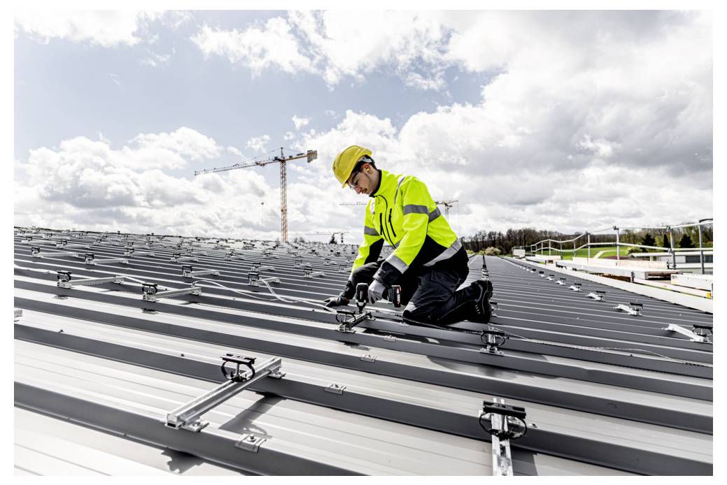 Bauarbeiter in Schutzhelm und Warnweste kniet auf einem Metalldach und befestigt Montageschienen für Solarmodule unter bewölktem Himmel.