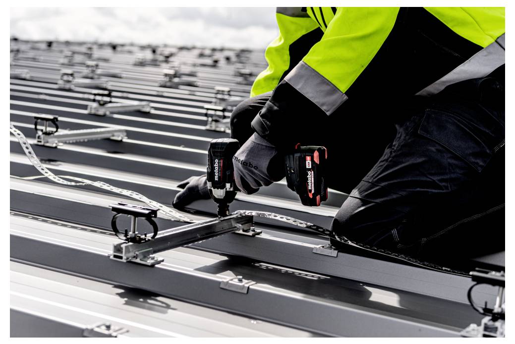 Arbeiter in Warnweste, der einen kabellosen Bohrer verwendet, um Metallbedachungspanele während der Dachinstallation zu befestigen.