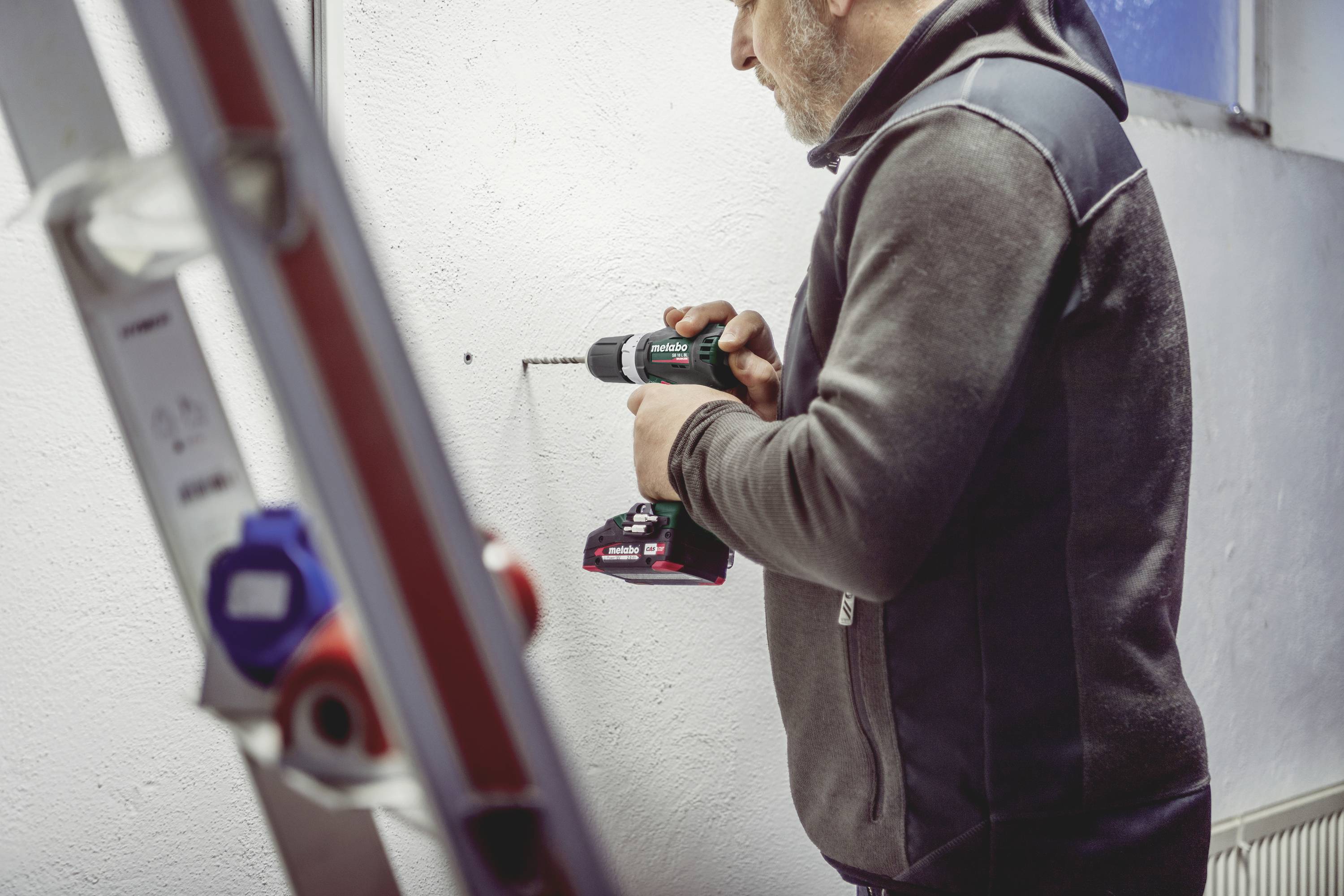 Ein Mann in Arbeitskleidung benutzt eine Bohrmaschine, um ein Loch in eine weiße Wand zu bohren. Eine Leiter ist im Vordergrund sichtbar.