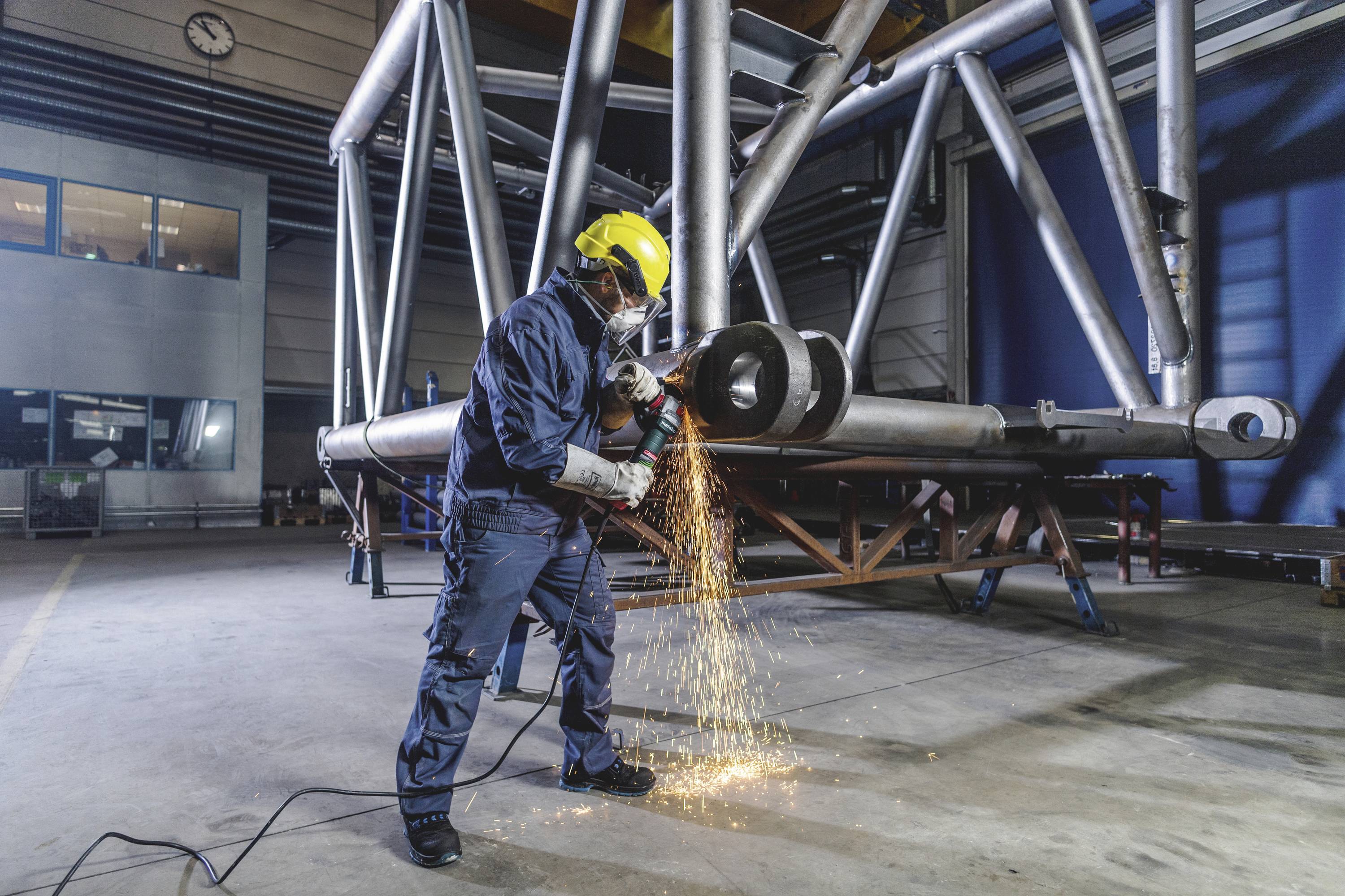 Ein Arbeiter in Schutzkleidung schleift Metallrohre in einer Fabrikhalle. Funken sprühen, während die Maschine das Metall bearbeitet.