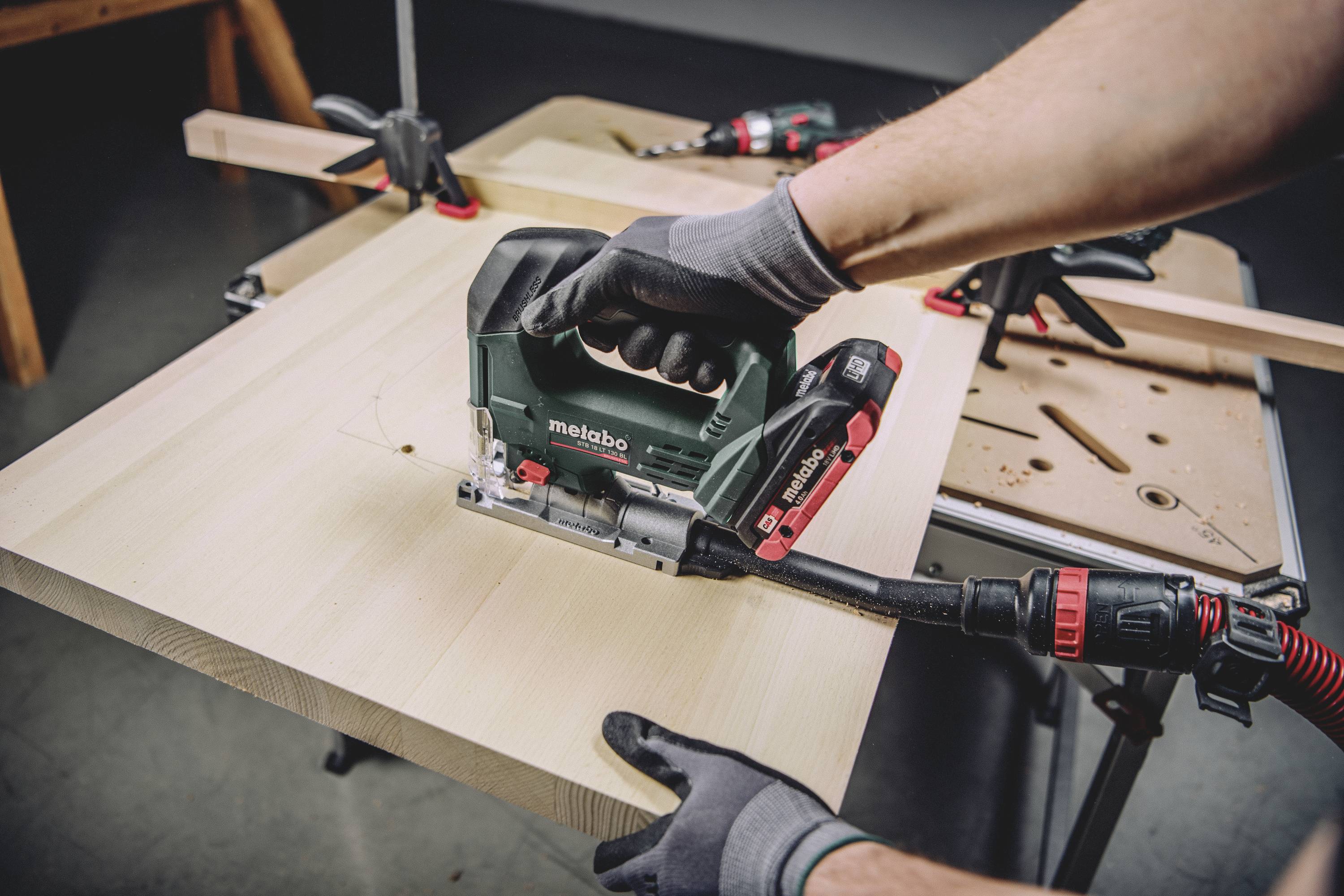 Eine Person sägt mit einer Metabo Stichsäge ein Holzbrett auf einem Arbeitstisch. Das Brett ist in einer Zwinge fixiert.