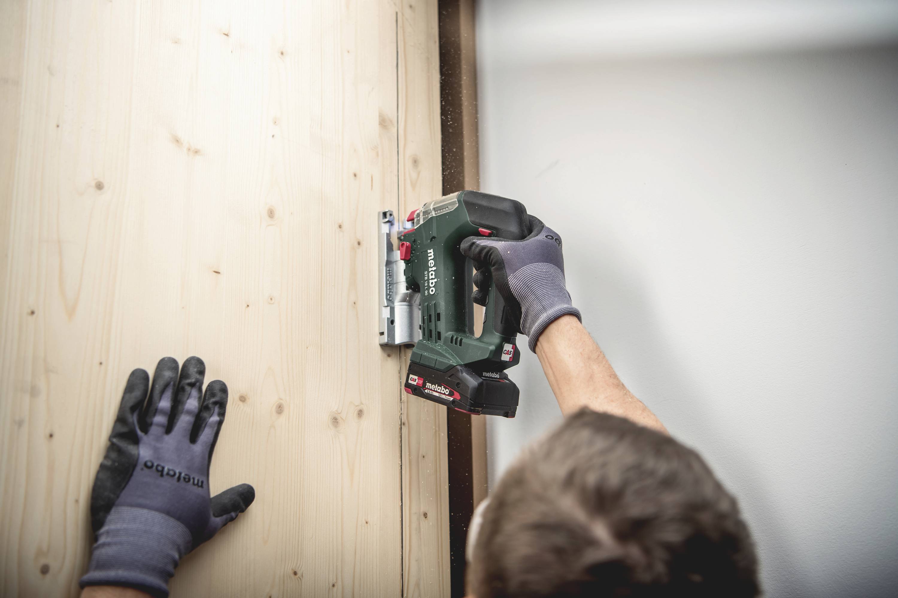 Eine Person benutzt einen Akkubohrer, um ein Holzbrett an eine Wand zu befestigen. Die Person trägt Arbeitshandschuhe.