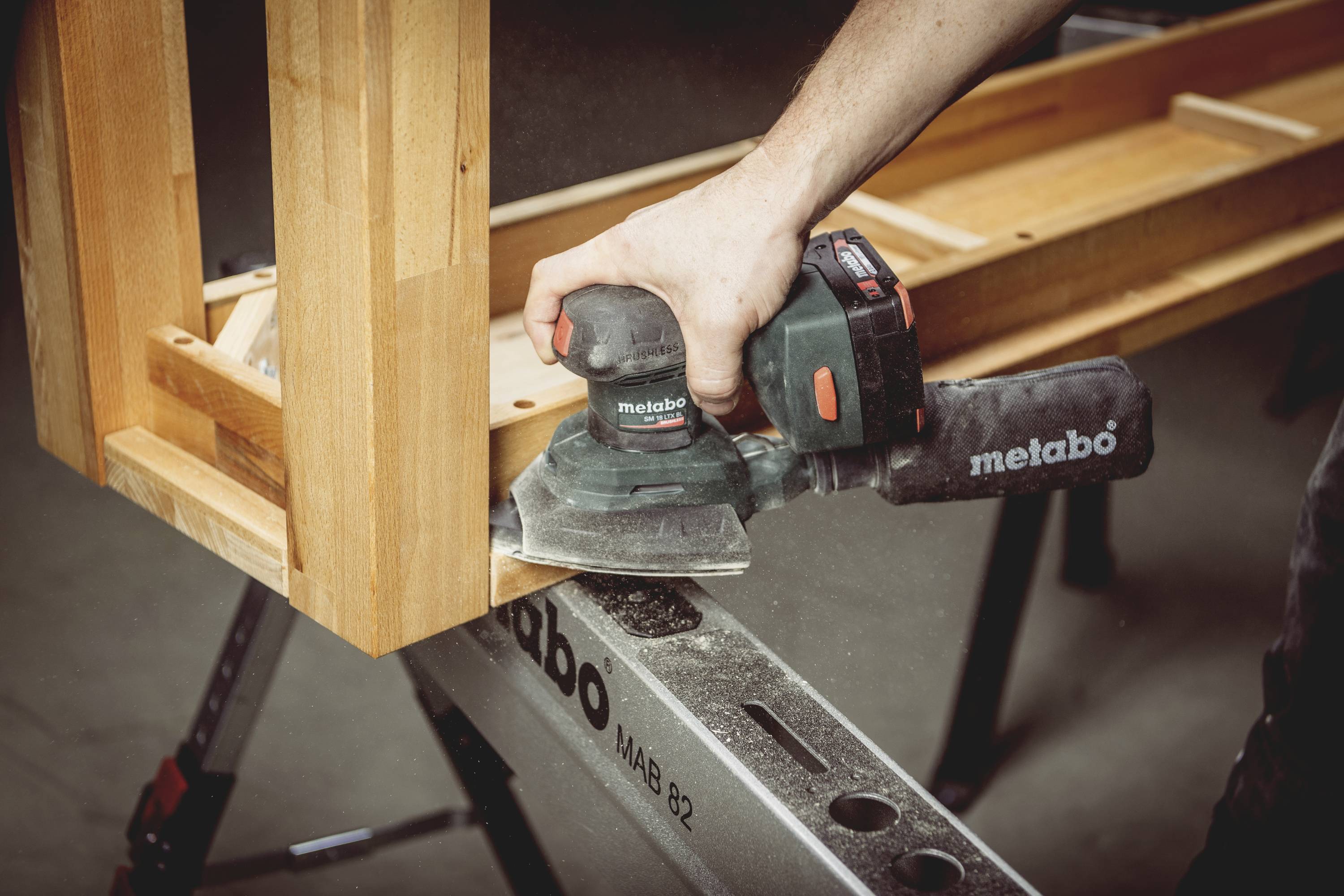 Eine Person schleift Holz mit einem Metabo Schleifgerät auf einer Werkbank. Holzspäne und Staub sind sichtbar.