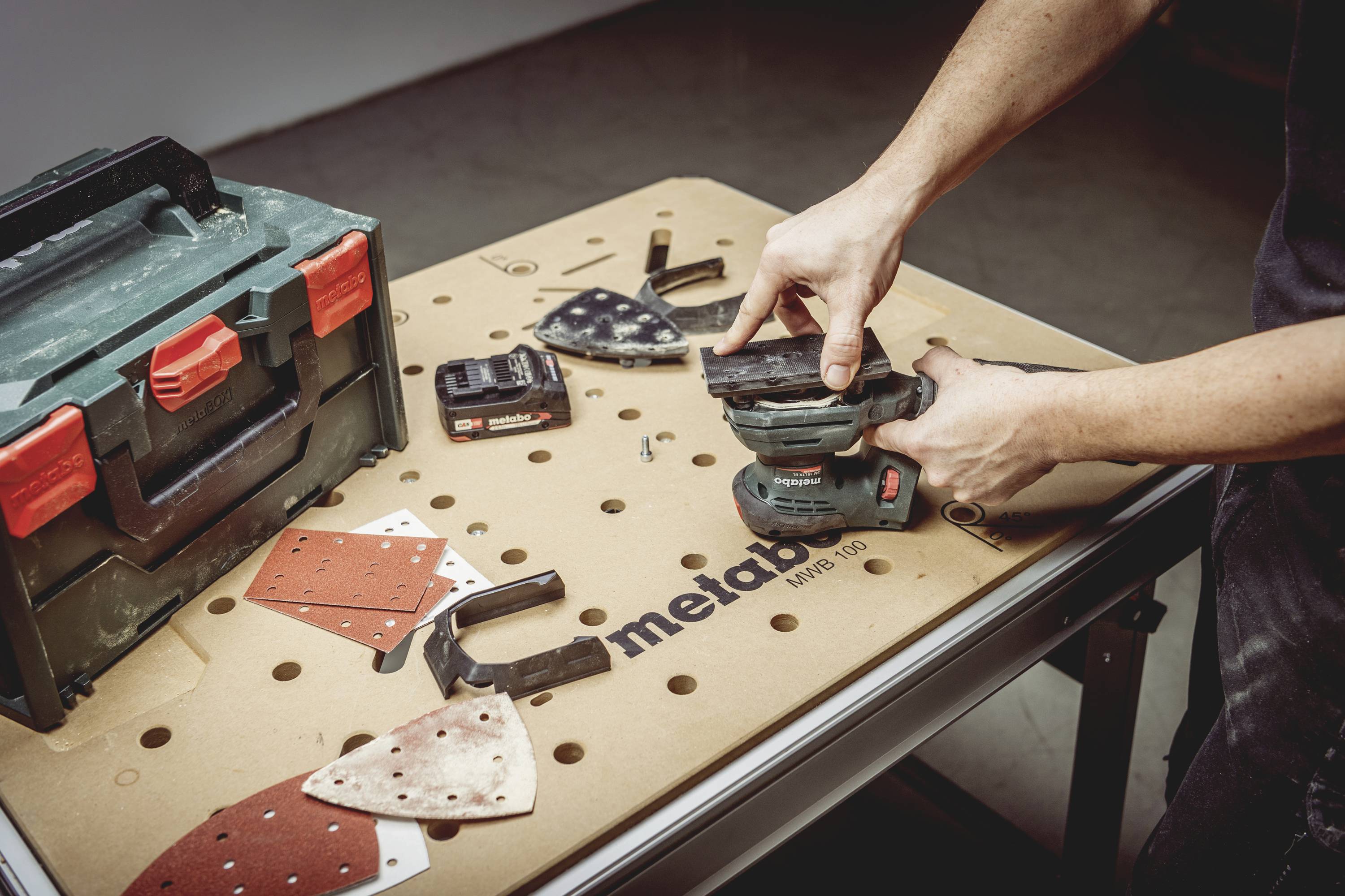 Ein Handwerker schleift ein Stück Holz auf einer Werkbank. Werkzeuge, Schleifpapier und eine Werkzeugkiste sind im Hintergrund zu sehen.