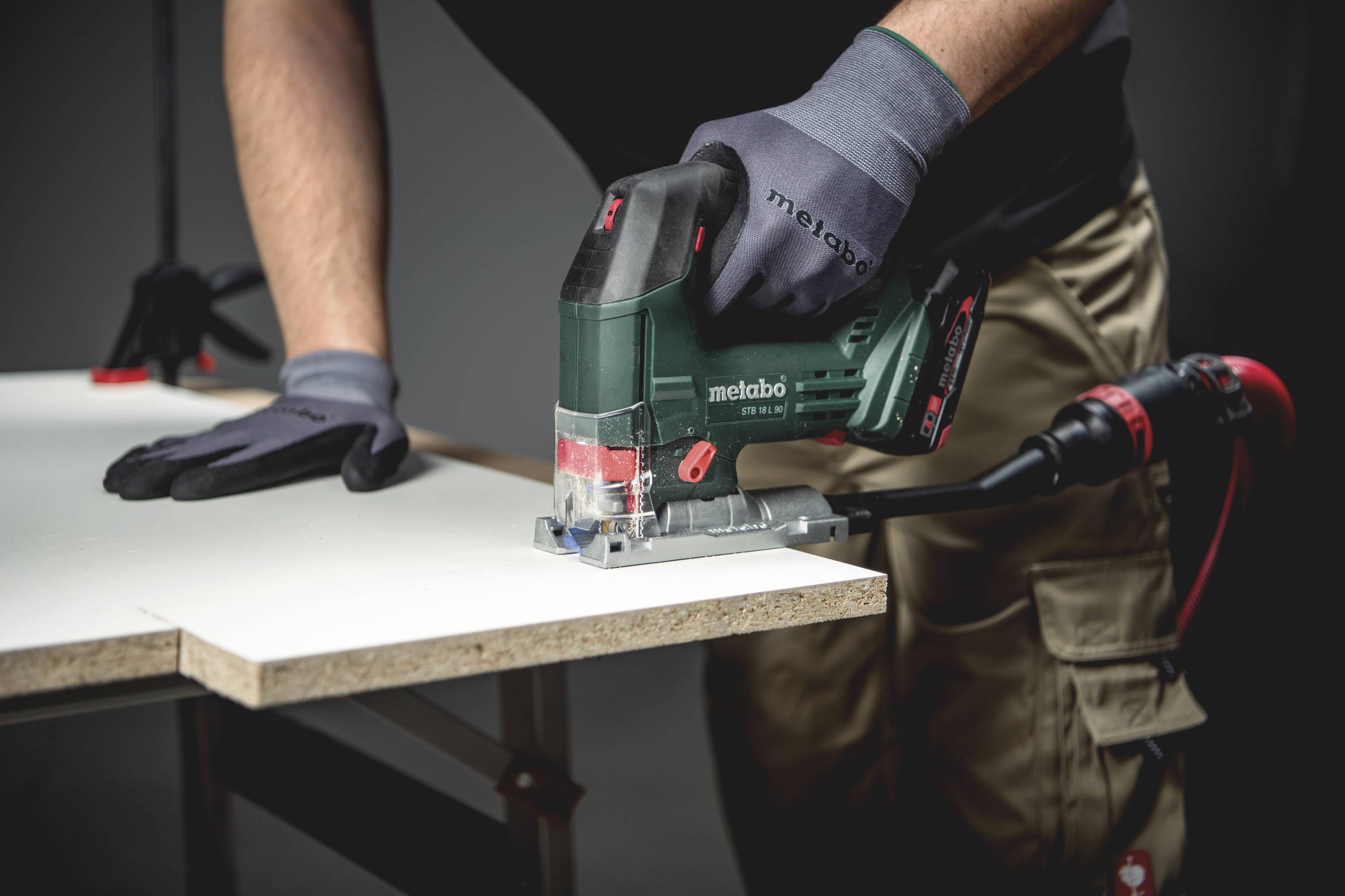 Eine Person schneidet mit einer Stichsäge eine weiße Holzplatte. Sie trägt Arbeitshandschuhe und eine dunkelgraue Arbeitskleidung.