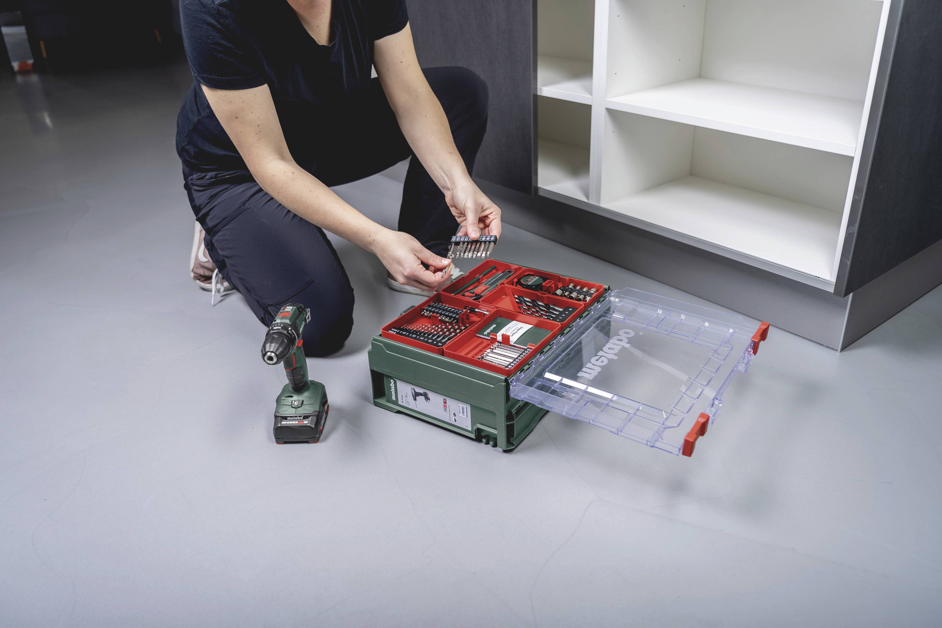 Eine Person kniet auf dem Boden und sortiert Schrauben in einer grünen Werkzeugbox neben einem Akkuschrauber und einem Regal.