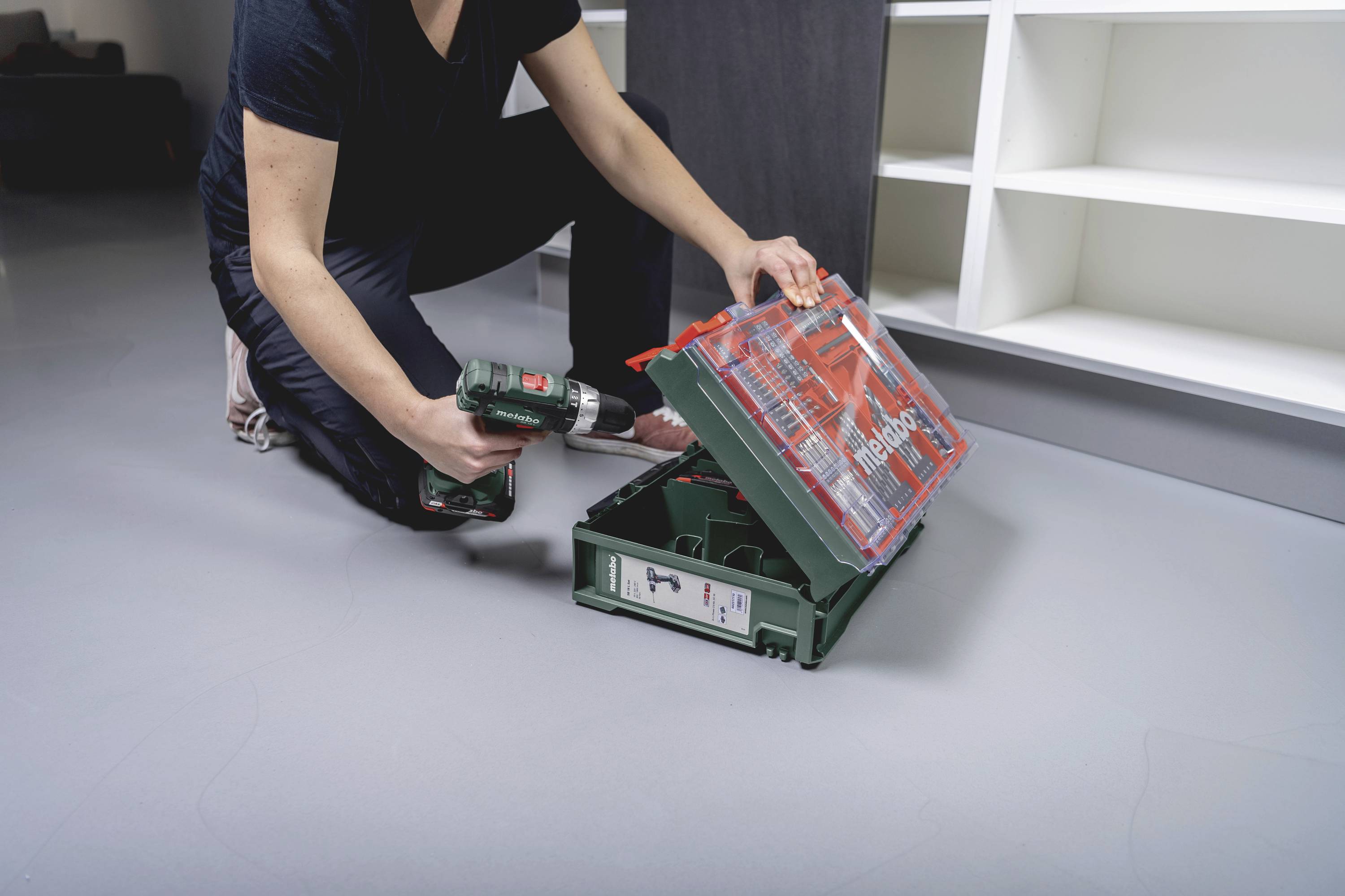 Eine Person kniet auf dem Boden und hält einen grünen Werkzeugkoffer mit einem Bohrer. Regal im Hintergrund.