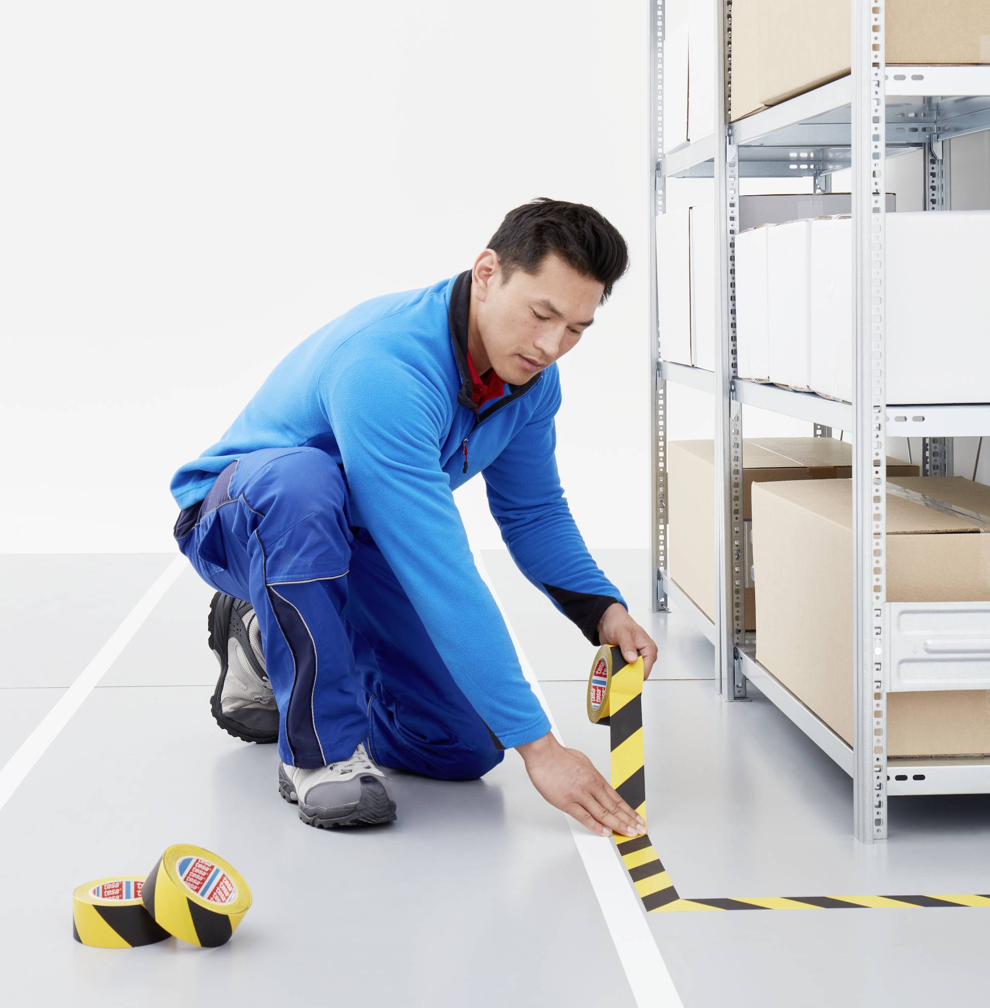 Ein Mann in blauer Arbeitskleidung klebt gelb-schwarzes Warnband auf den Boden eines Lagers neben Regalen mit Kartons.