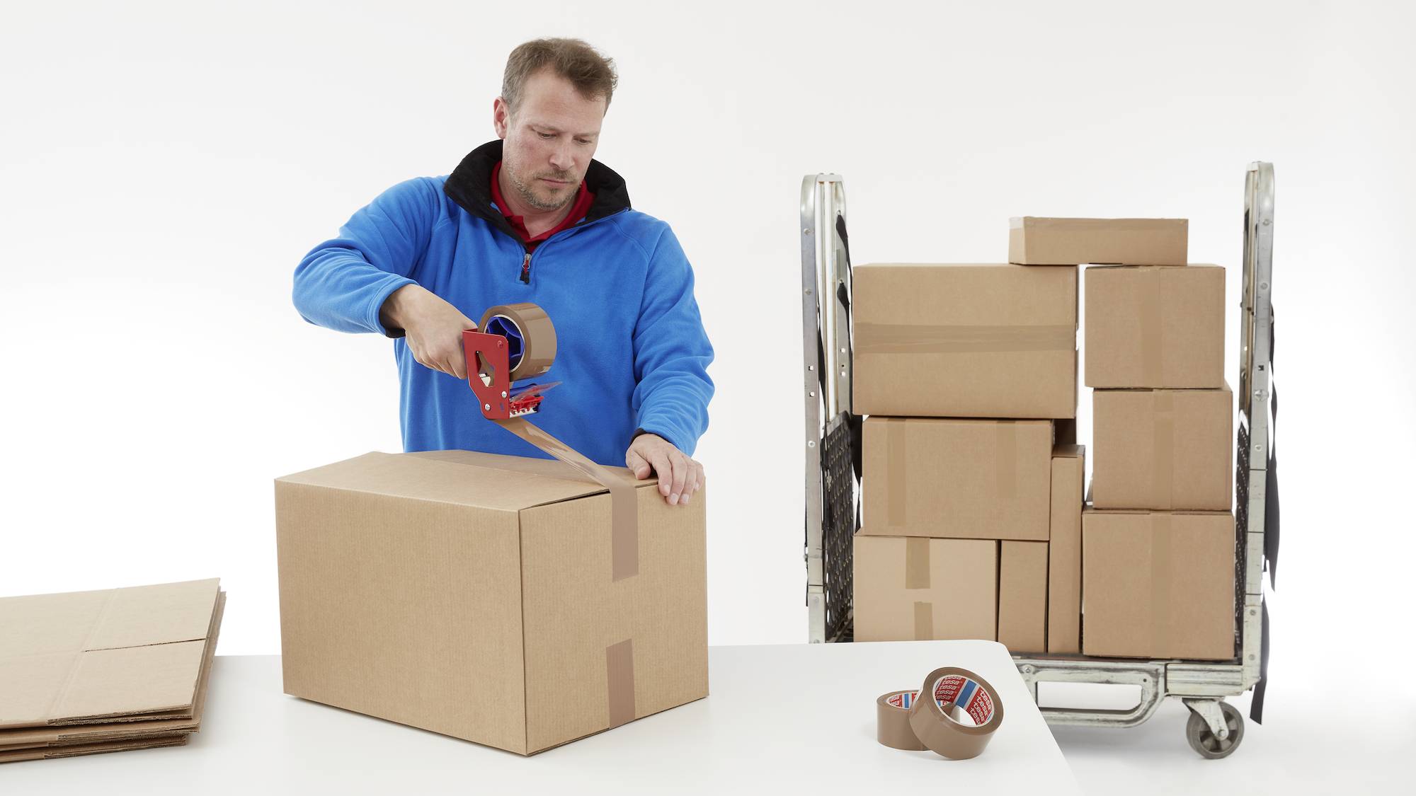 Ein Mann verpackt einen Karton mit Klebeband; im Hintergrund sind weitere Kartons auf einem Rollwagen gestapelt.