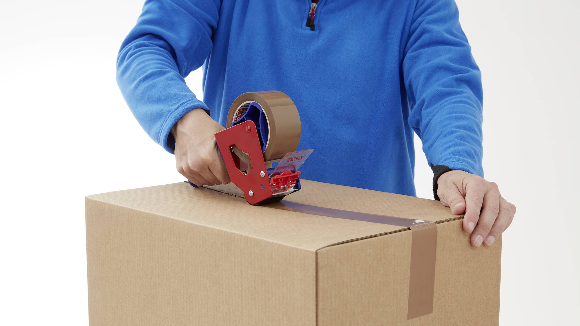 Person in blauem Pullover versiegelt einen Karton mit Klebeband.