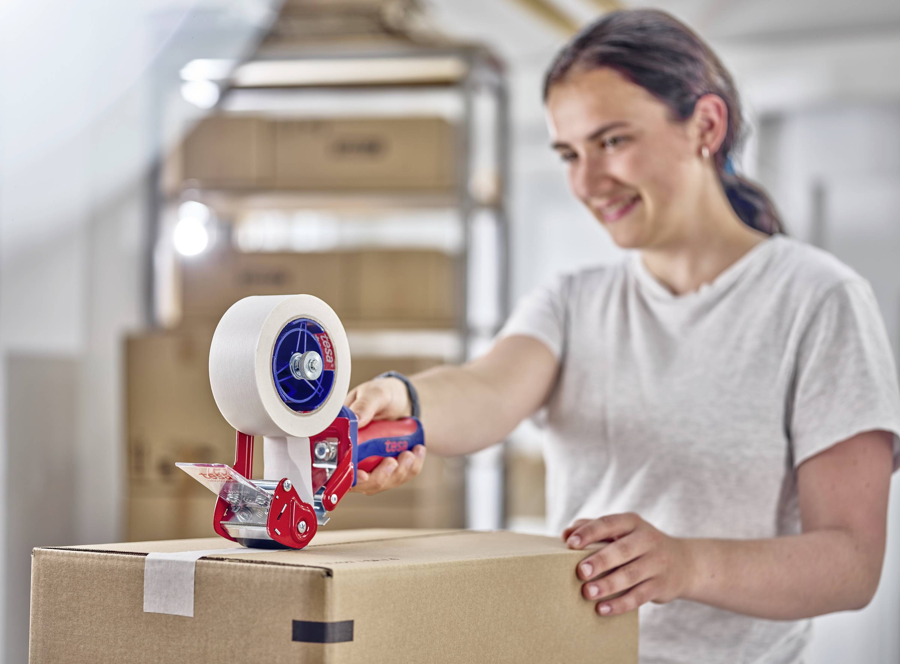 Eine lächelnde Person verpackt einen Karton mit einem Klebebandabroller in einem Raum mit Kartons im Hintergrund.