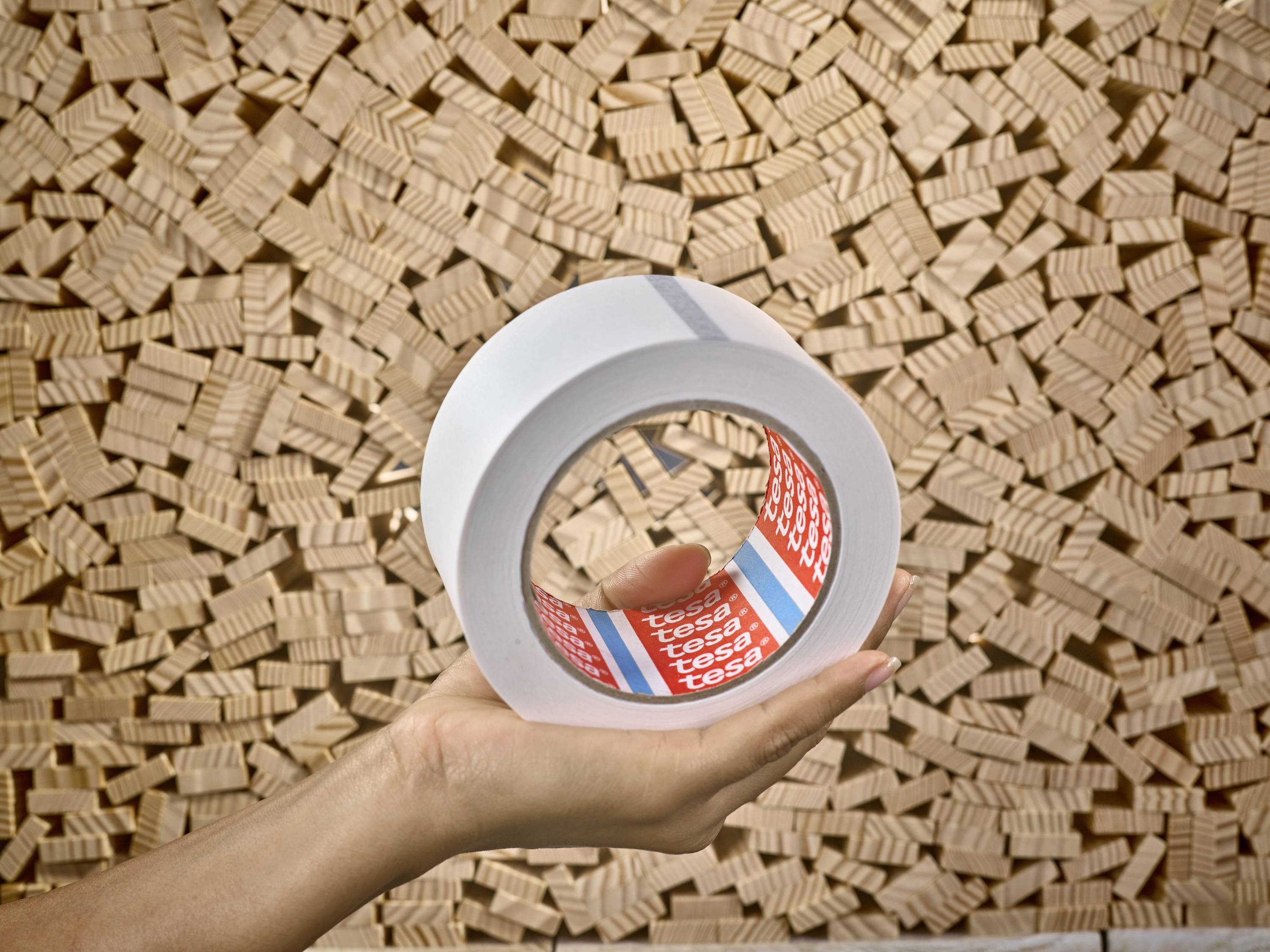 Eine Hand hält eine große Rolle durchsichtiges Klebeband vor einem Hintergrund aus gestapelten Holzwürfeln.