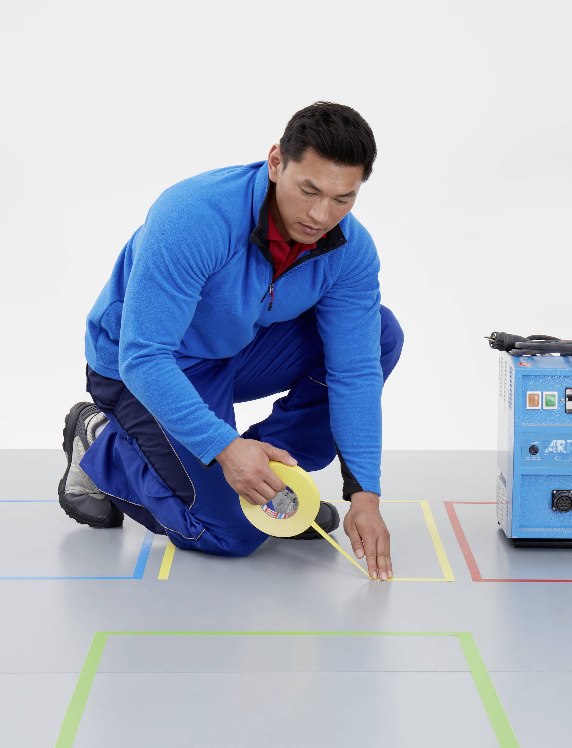 Eine Person kniet auf einem grauen Boden und klebt gelbes Klebeband in rechteckigen Mustern. Neben ihr steht ein blaues Gerät.