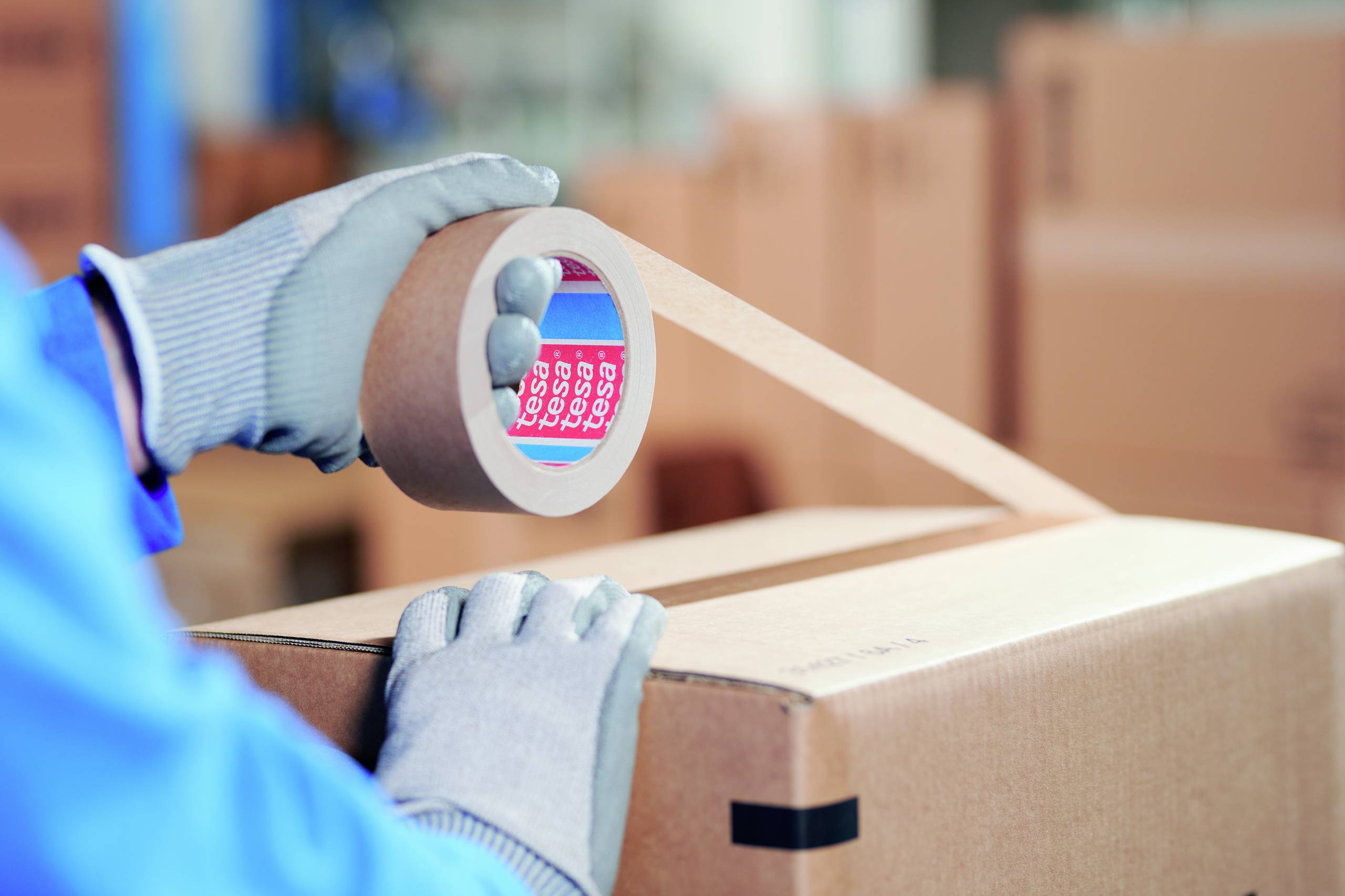 Person in blauer Kleidung und Handschuhen klebt einen Karton mit Klebeband zu. Der Karton befindet sich in einer Umgebung voller Packkartons.