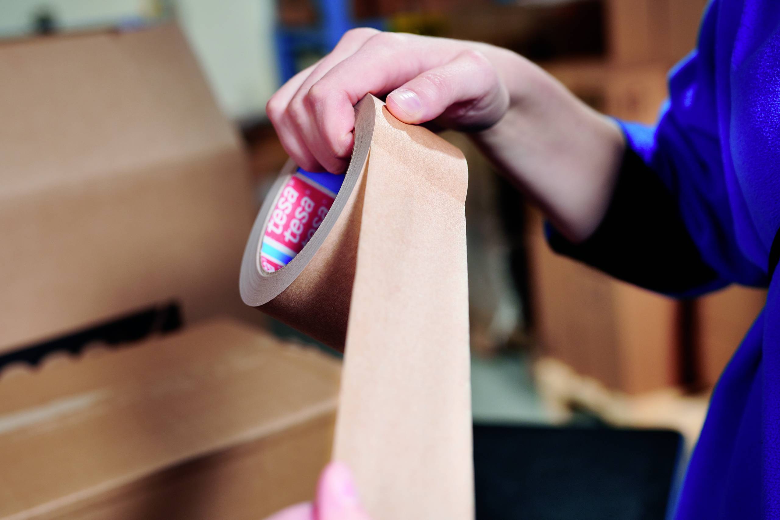 Eine Person hält eine Rolle braunes Klebeband und zieht ein Stück davon ab; im Hintergrund steht ein offener Karton.