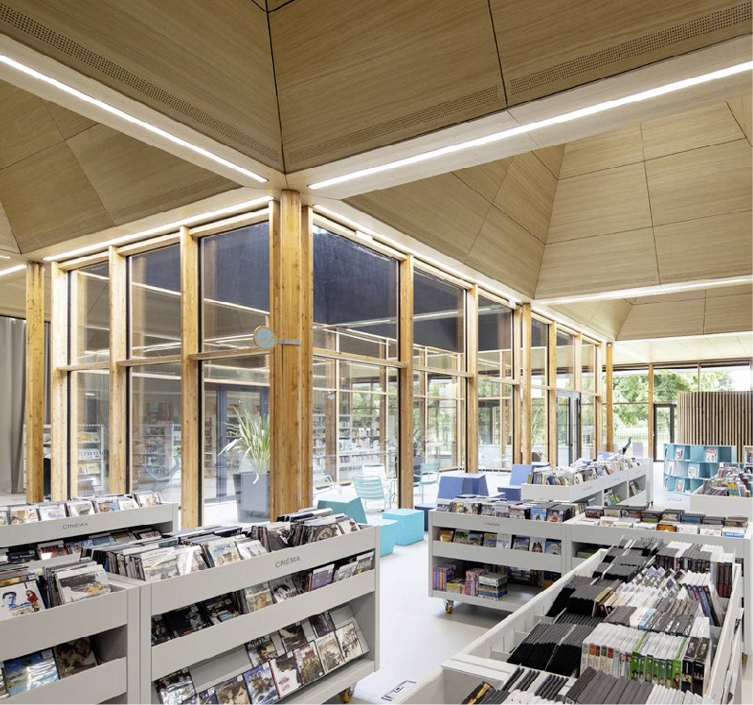 Ein heller, moderner Bibliotheksraum mit Holzakzenten. Regale sind mit Büchern und Medien gefüllt. Große Fenster bieten Tageslicht.