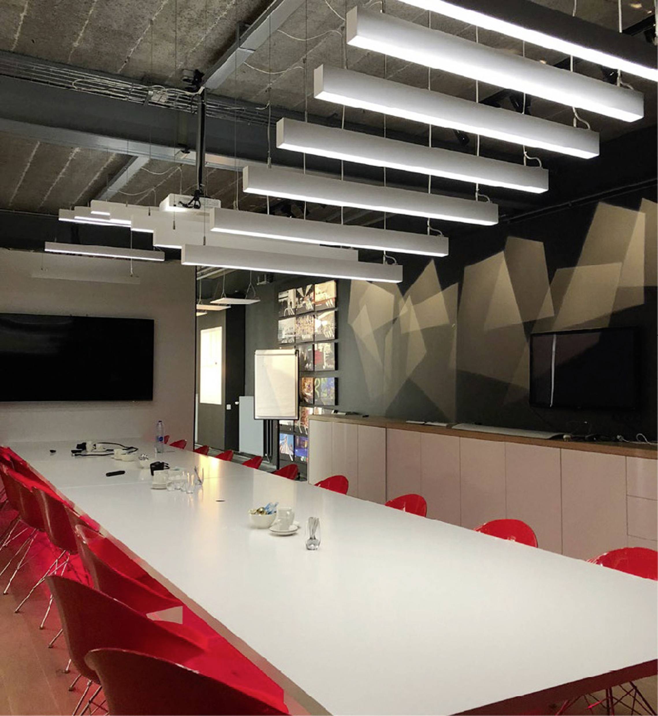 Konferenzraum mit großem weißen Tisch, roten Stühlen, an der Decke hängenden Leuchten, und einem großen Bildschirm an der Wand.