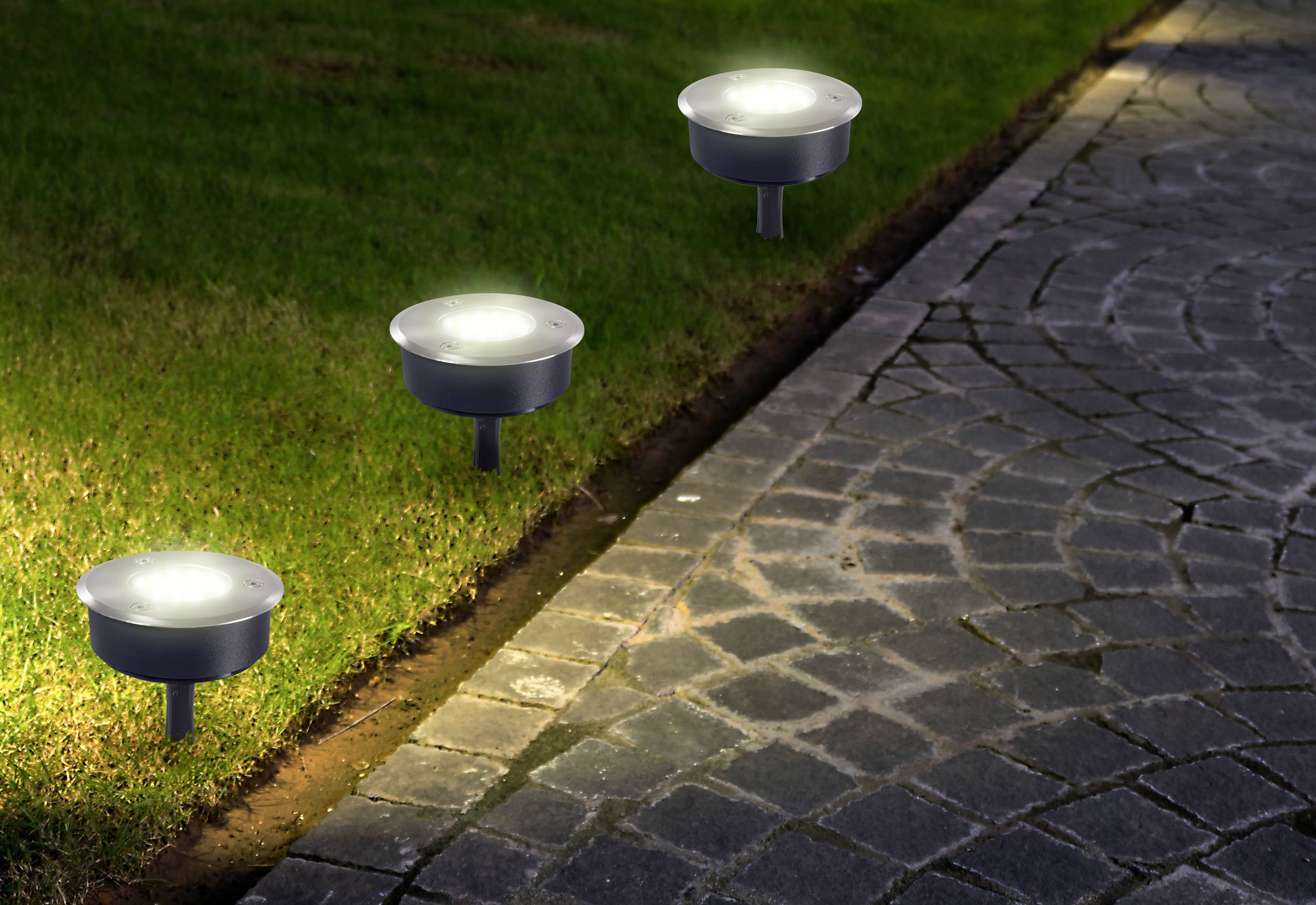 Solarbetriebene Wegeleuchten beleuchten nachts ein Grasfläche neben einem gepflasterten Gehweg.