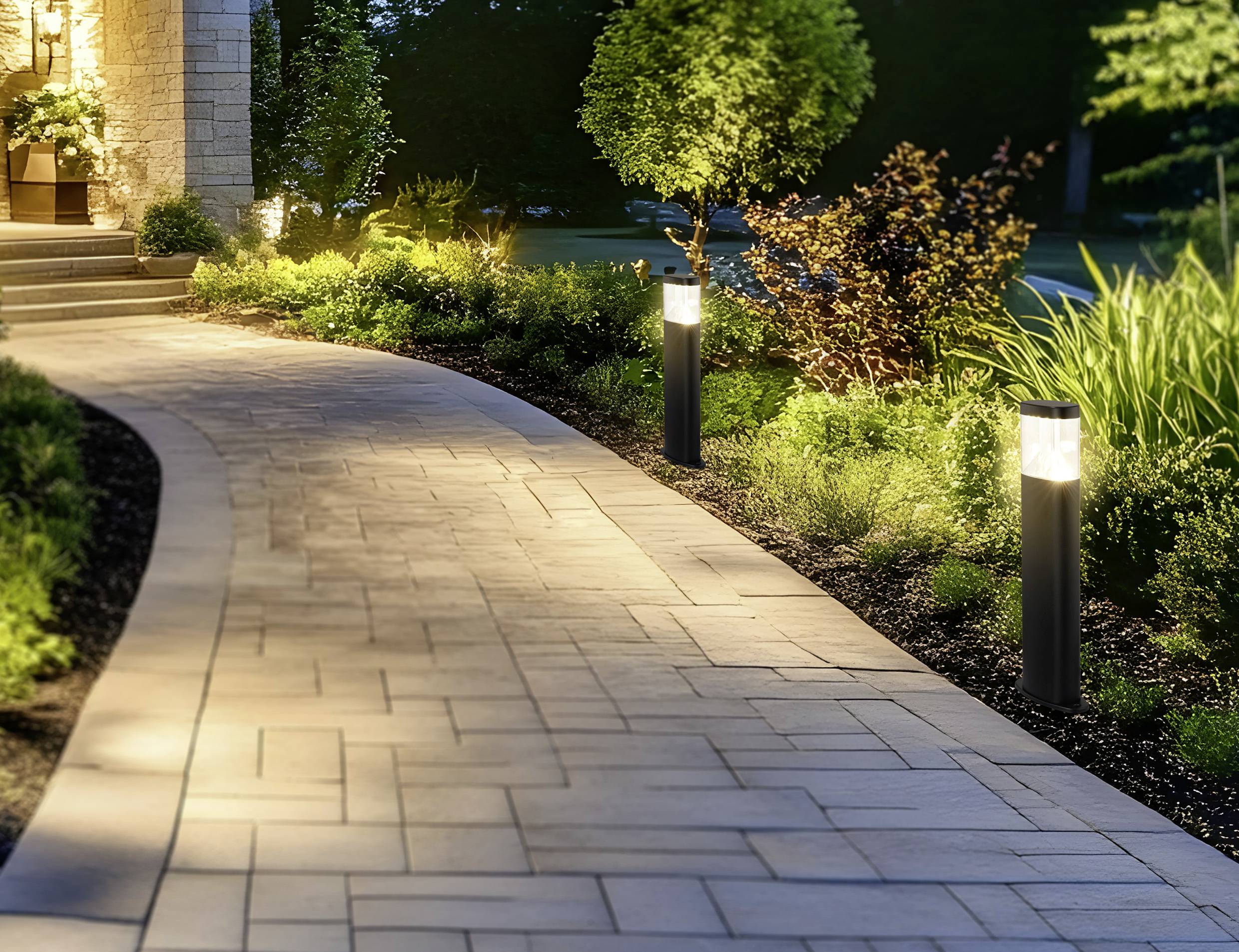 Ein geschwungener Steinweg, gesäumt von Gartenleuchten, führt durch eine landschaftlich gestaltete Fläche mit üppigem Grün und Sträuchern zum Hauseingang in der Abenddämmerung.