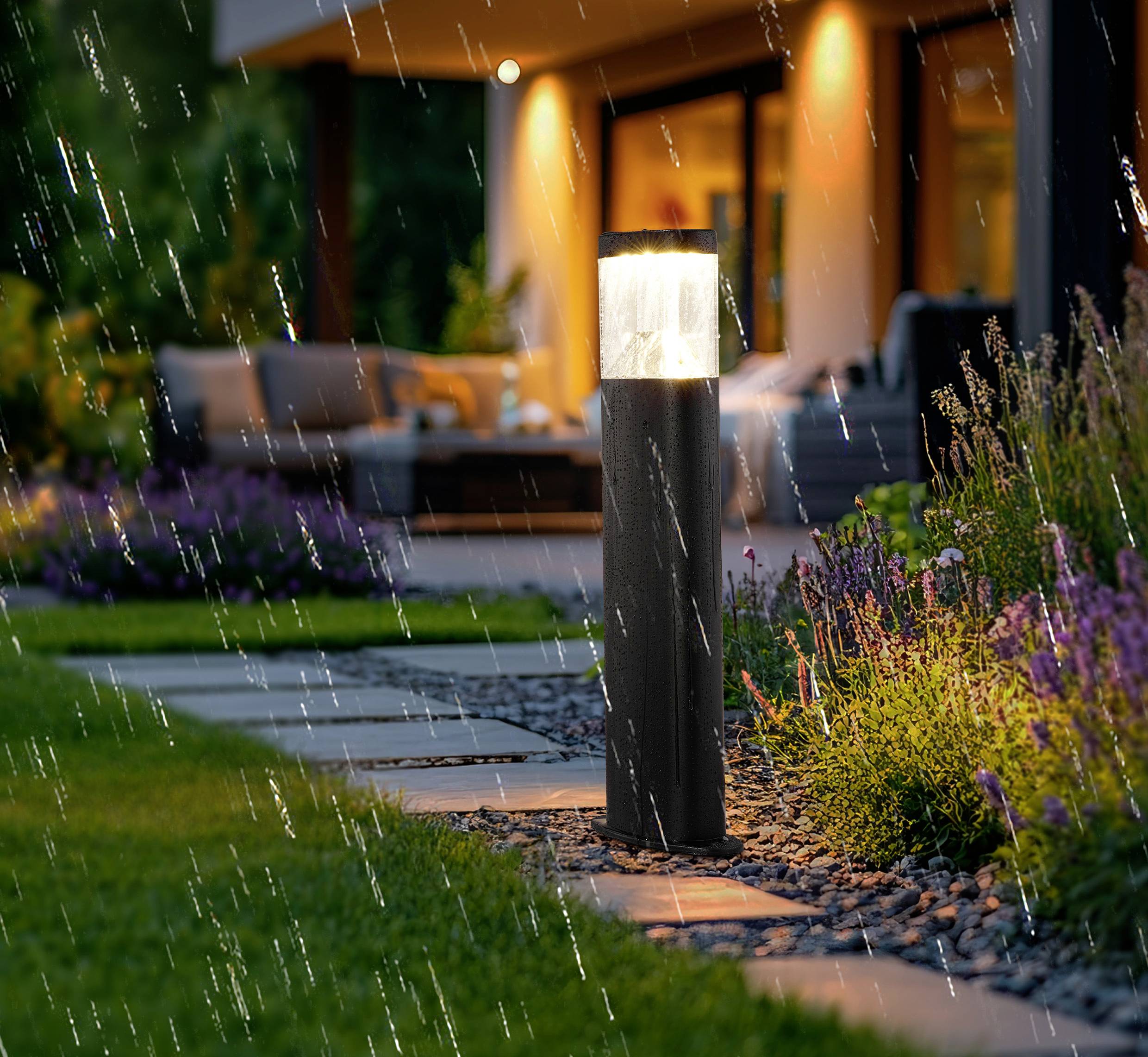 Moderner Außengarten mit einem leuchtenden Wegbeleuchtungselement, umgeben von feuchtem Laub und Regen, der zu einem warm beleuchteten Haus führt.