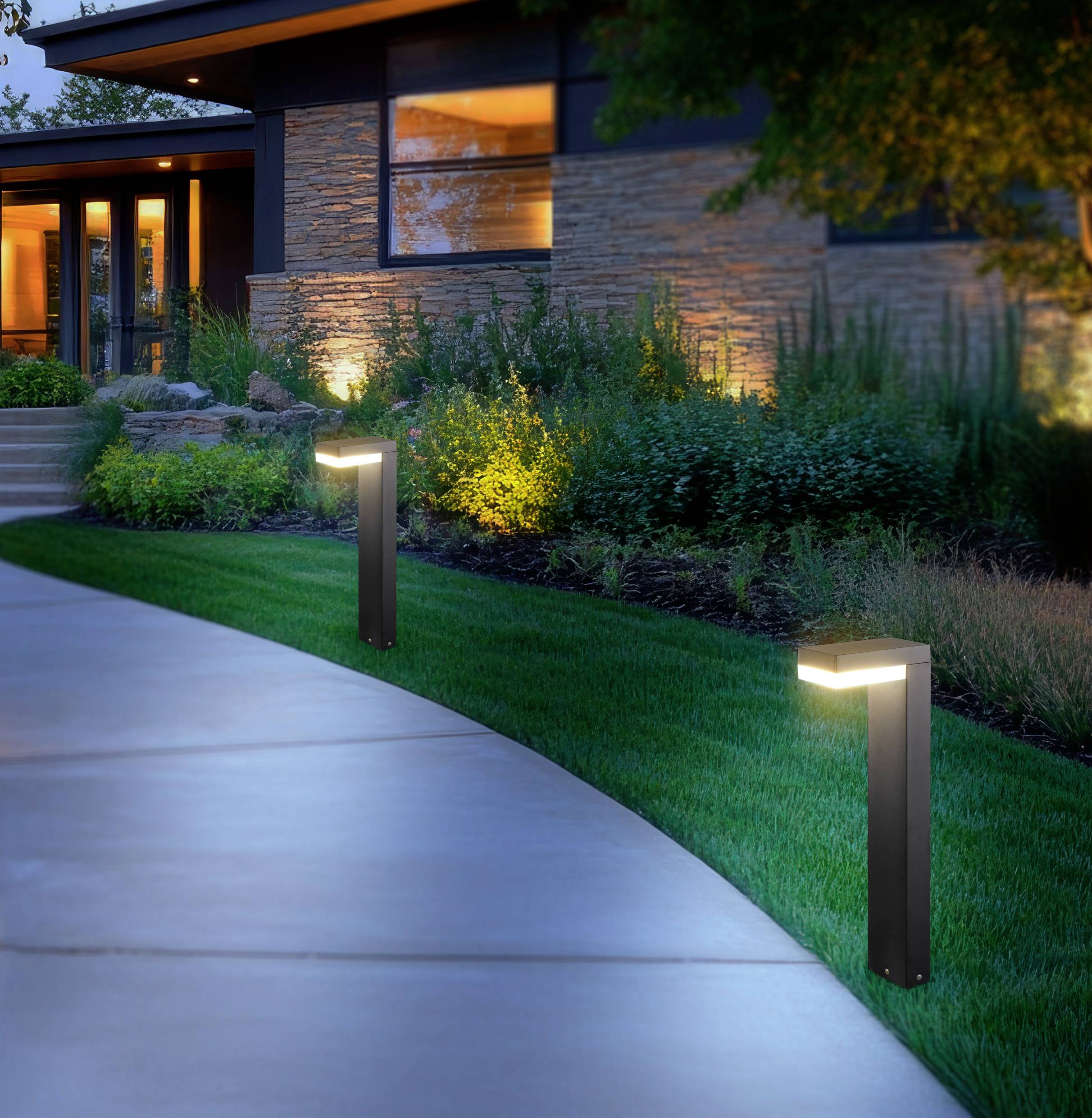 Ein modernes Haus mit Außenbeleuchtung. Zwei schlanke, beleuchtete Wegelampen säumen einen geschwungenen Gehweg, der von gepflegtem Rasen und Sträuchern umgeben ist, kurz vor Einbruch der Dunkelheit.