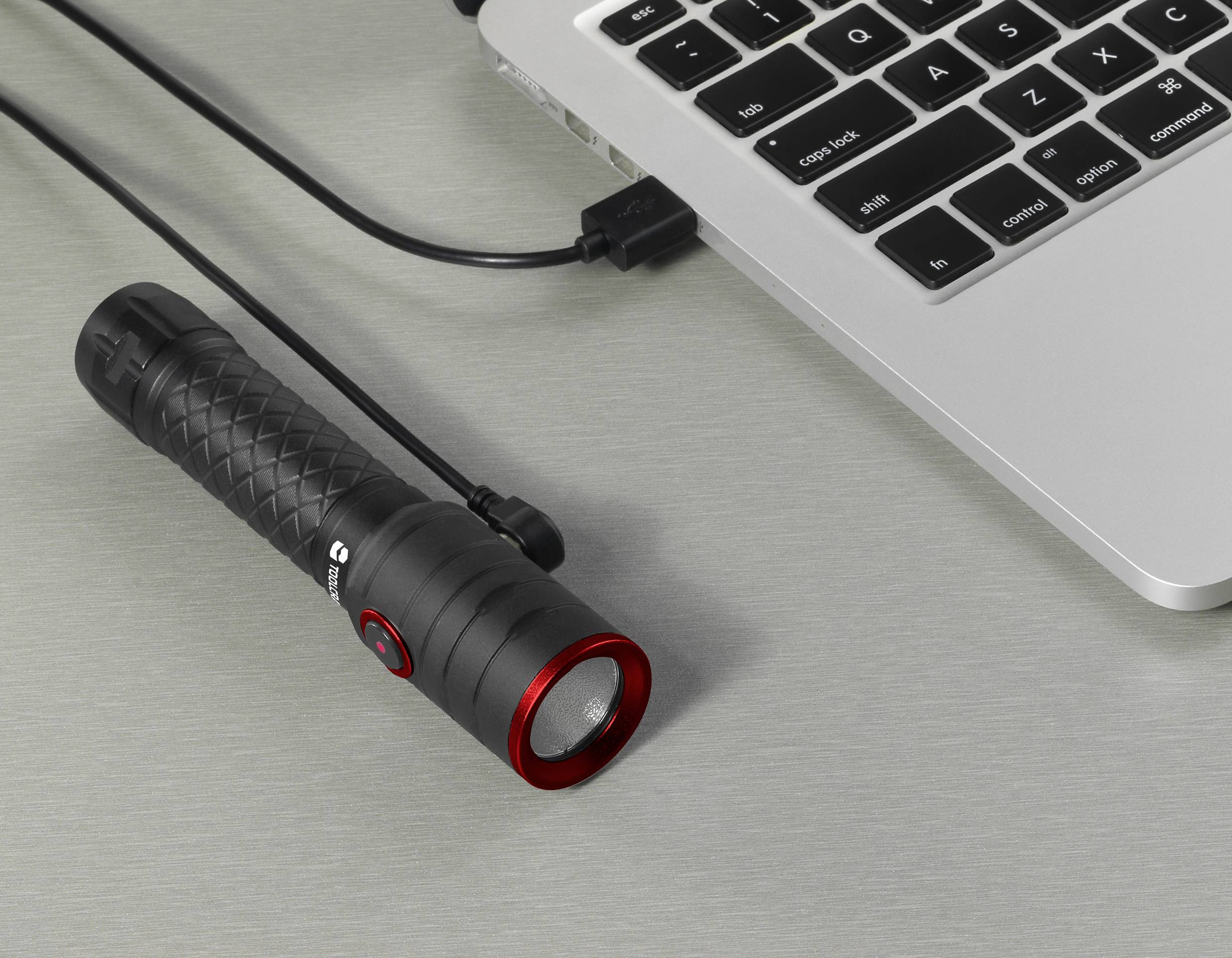 Eine schwarze Taschenlampe mit rotem Rand ist über ein USB-Kabel mit einem Laptop verbunden. Sie liegt auf einem Tisch.