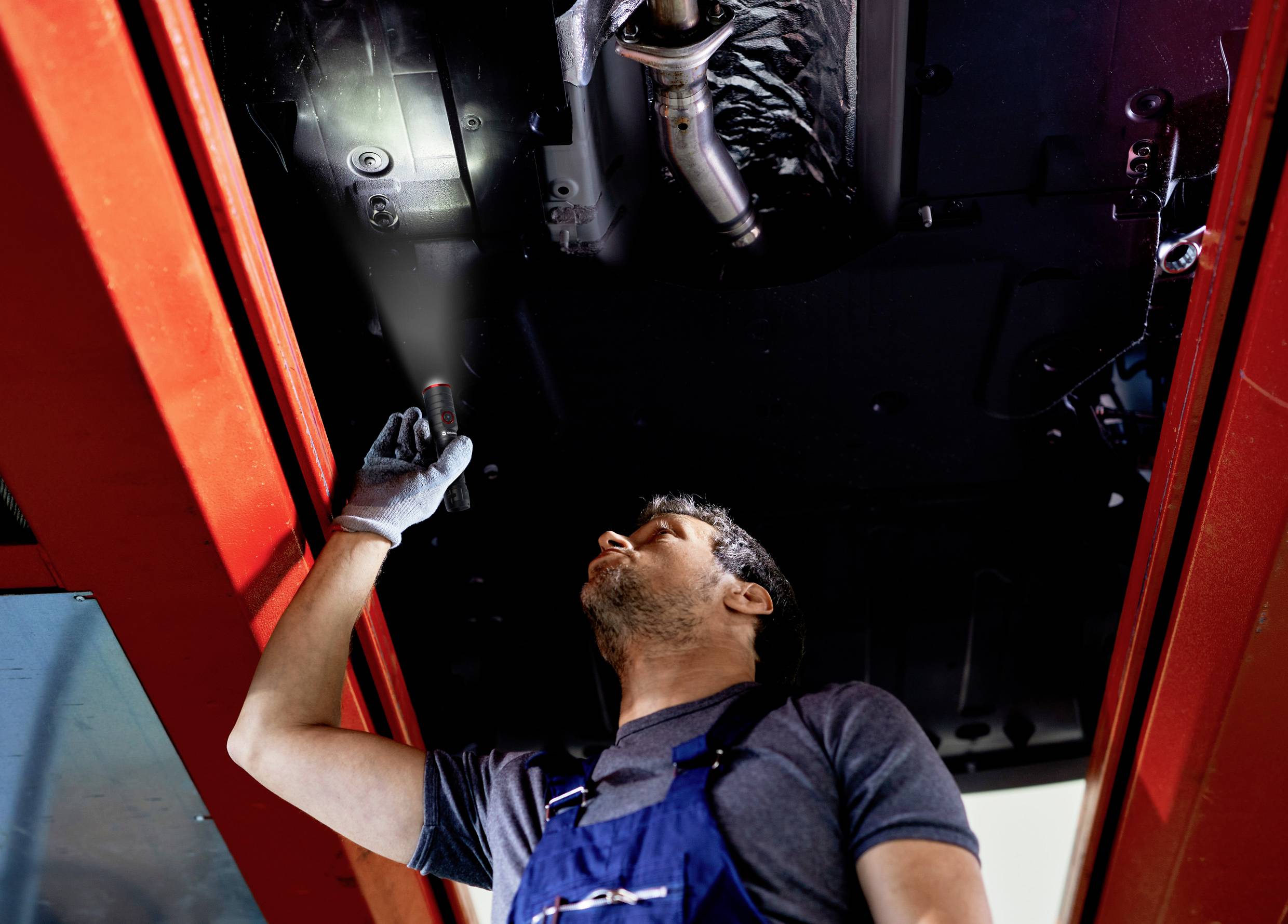 Ein Mechaniker in blauer Latzhose inspiziert den Unterboden eines Fahrzeugs in einer Werkstatt, während er mit einer Taschenlampe leuchtet.