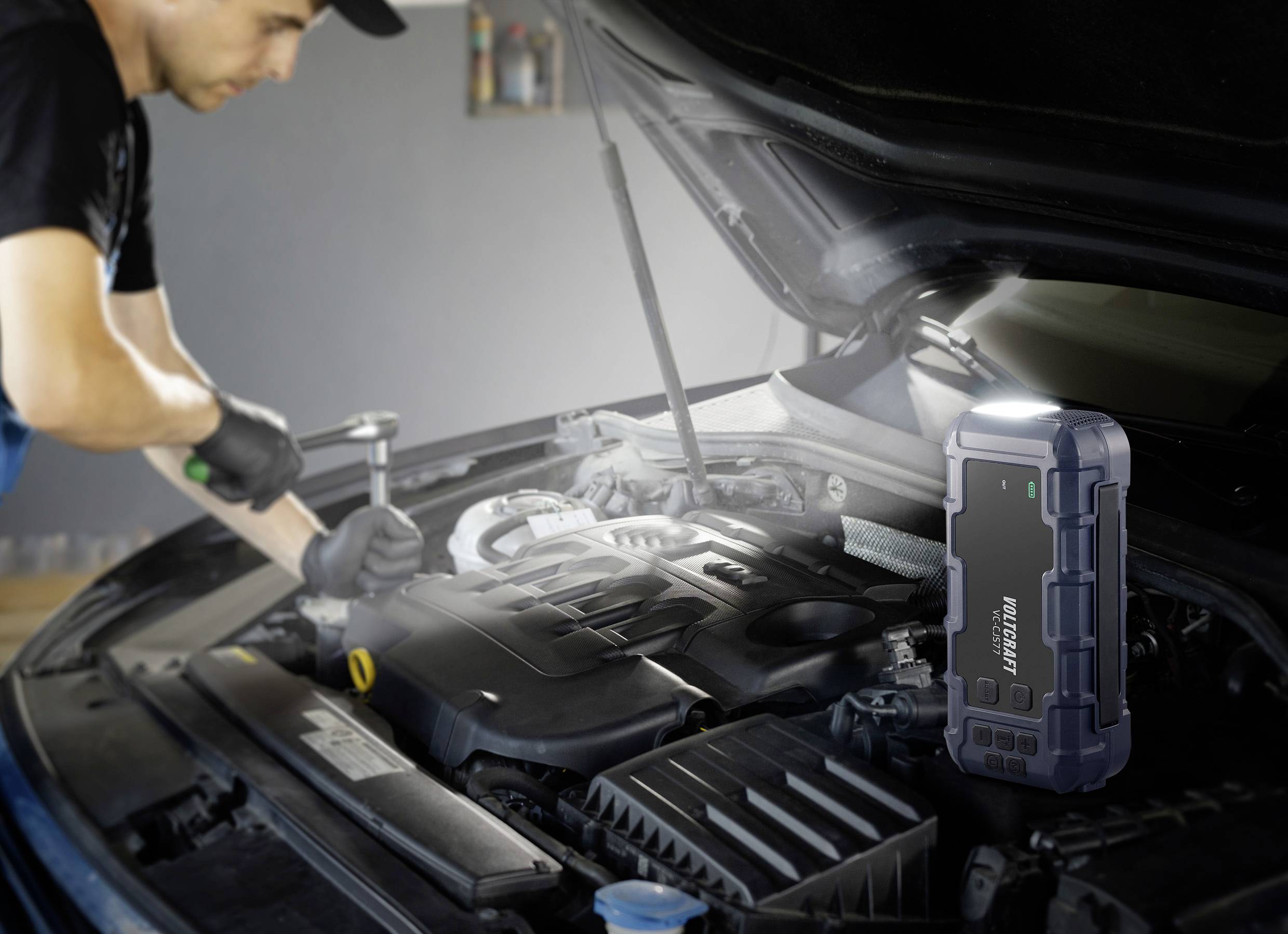 Ein Mechaniker arbeitet am Motor eines Autos mit geöffnetem Motorraum. Im Vordergrund steht ein tragbares Starthilfegerät.