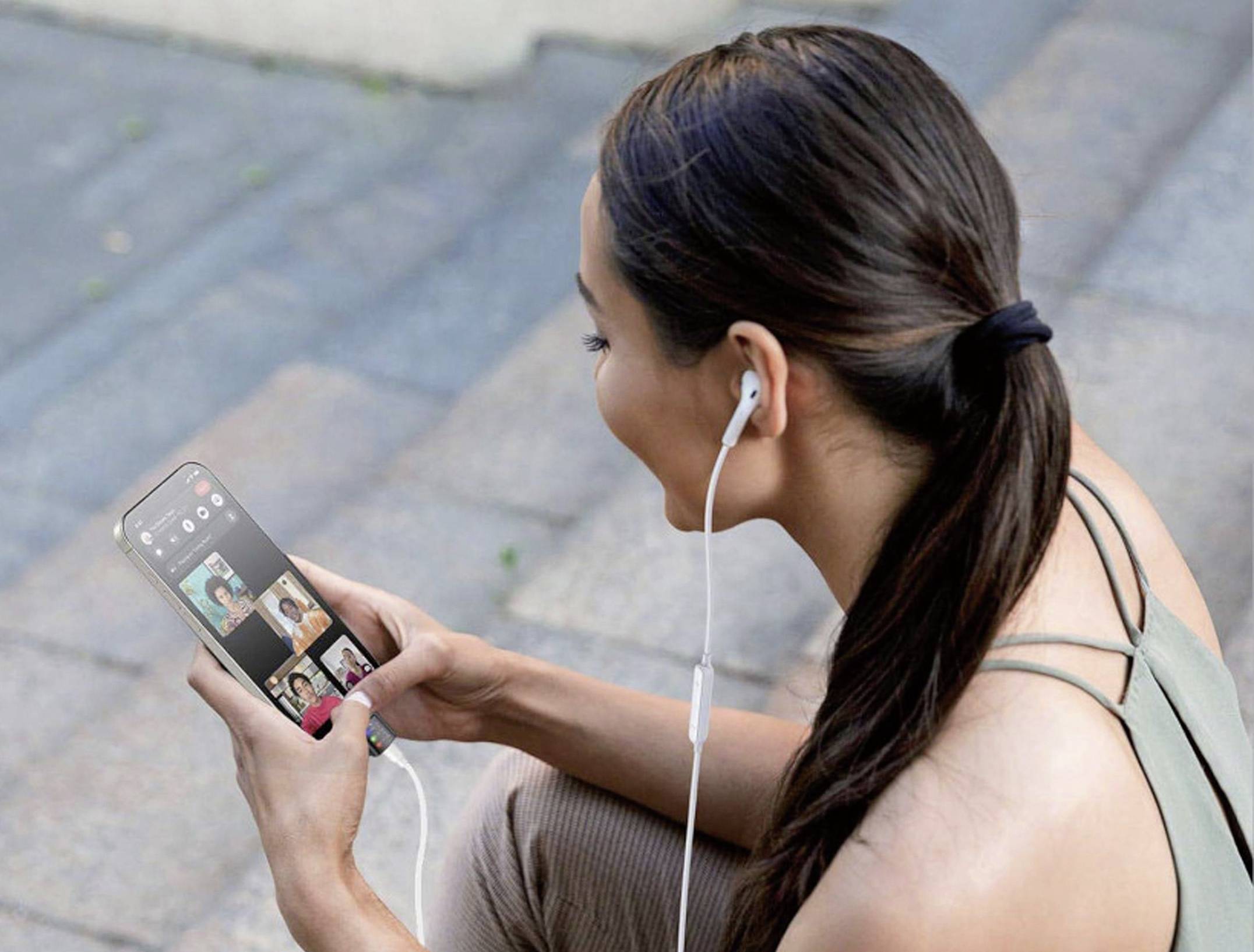 Frau sitzt draußen und nutzt ihr Smartphone für einen Videoanruf mit vier Personen. Sie trägt Ohrhörer und lächelt.