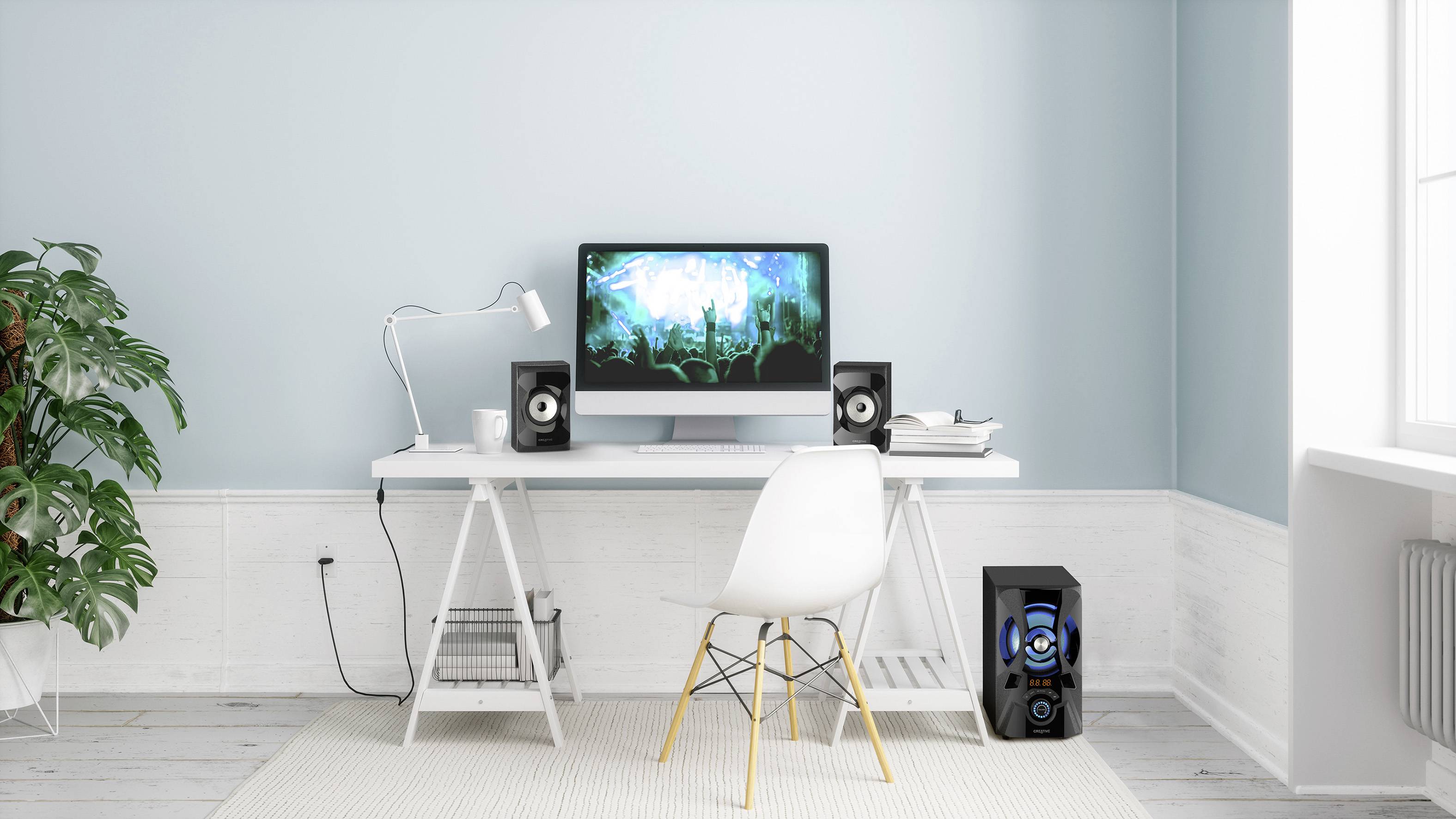 Ein moderner Schreibtisch mit Computer, zwei Lautsprechern, Schreibtischlampe und Büchern. Im Hintergrund ist eine Pflanze sichtbar.