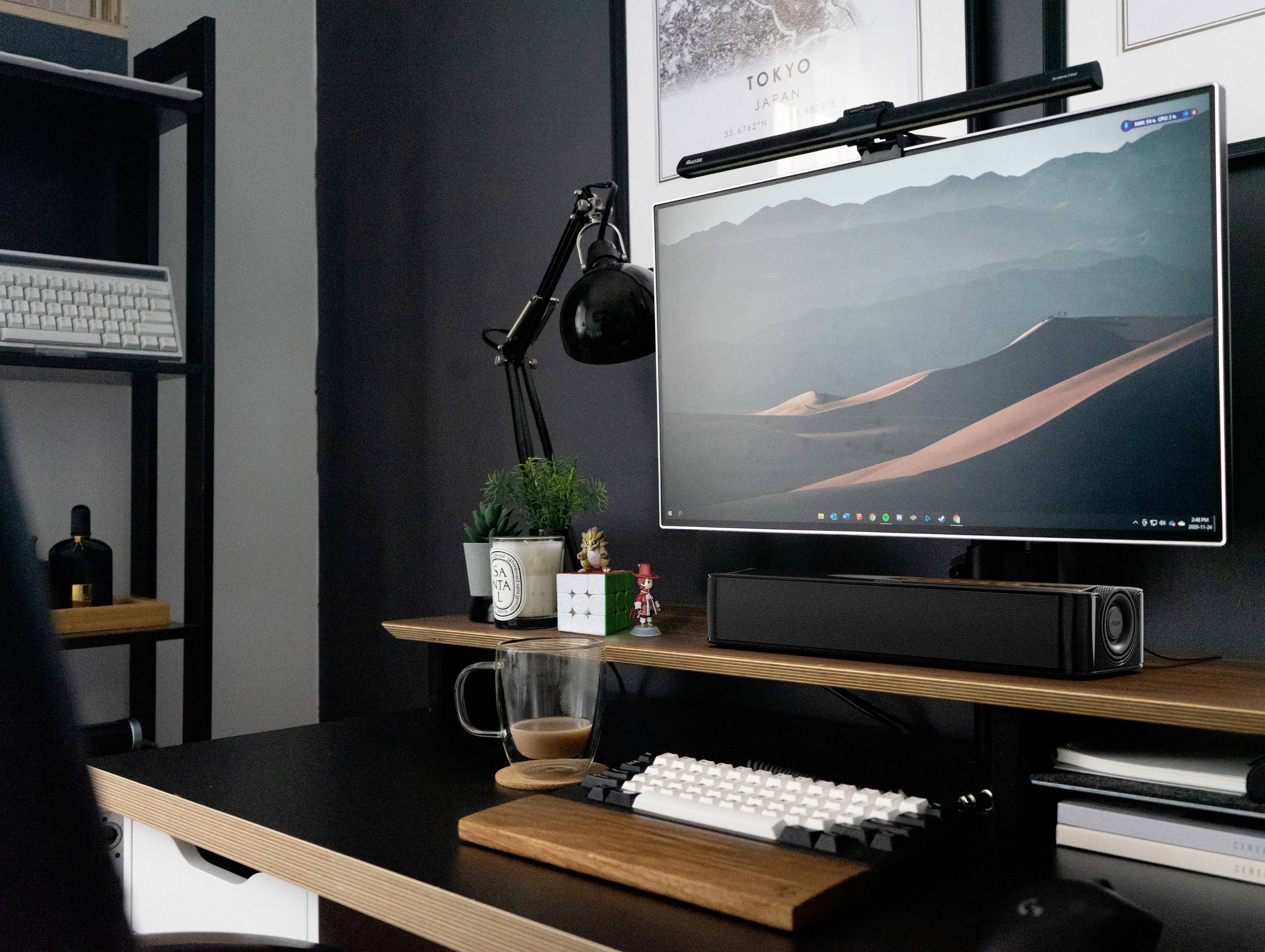 Moderner Schreibtisch mit Bildschirm zeigt Wüstenlandschaft. Tastatur, Lampe, Lautsprecher und Pflanzen arrangiert für kreative Arbeit.