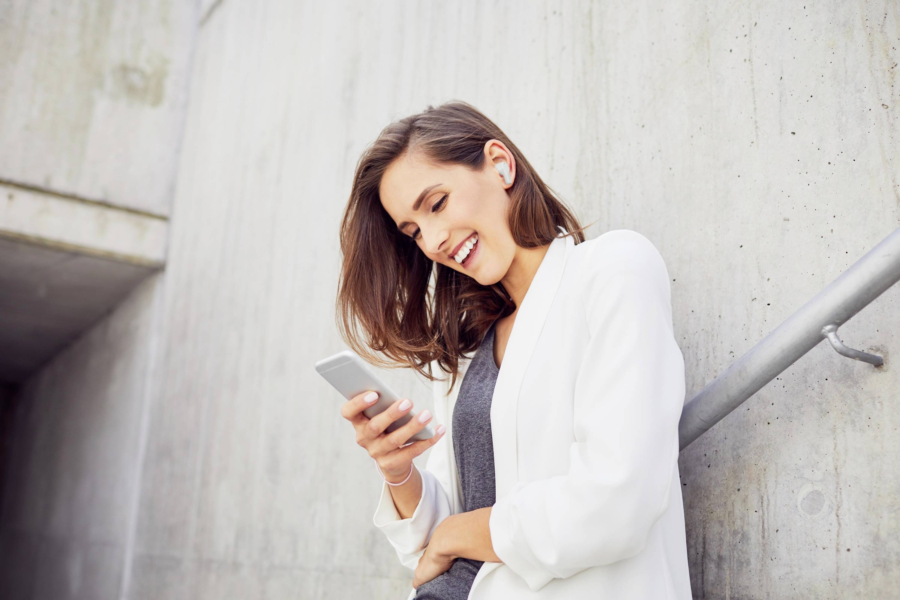 Eine Frau lehnt lächelnd an einer Betonwand und schaut auf ihr Smartphone. Sie trägt Kopfhörer und eine weiße Jacke.