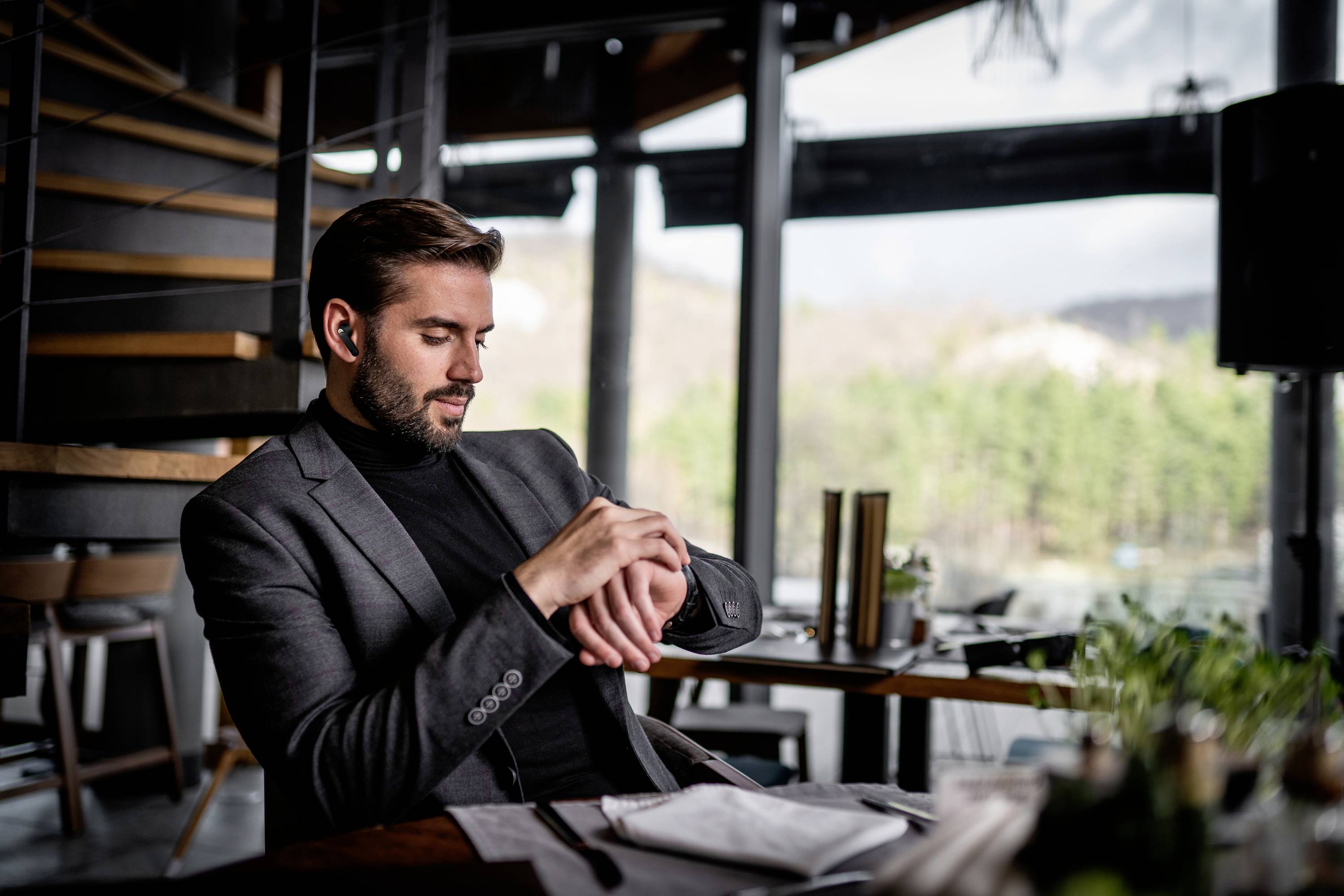 Ein Mann in einem Anzug sitzt in einem modernen Restaurant am Tisch und schaut auf seine Armbanduhr. Im Hintergrund ist Natur zu sehen.