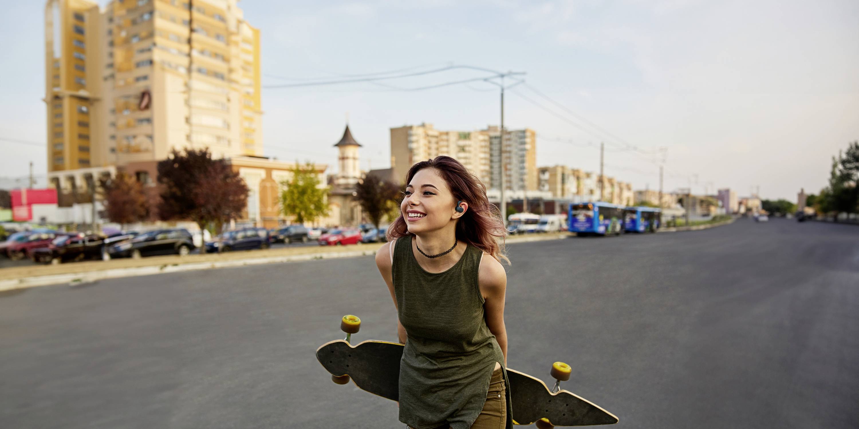 Eine lachende Frau hält ein Longboard und läuft auf einer breiten Straße bei sonnigem Wetter. Im Hintergrund sind Gebäude zu sehen.