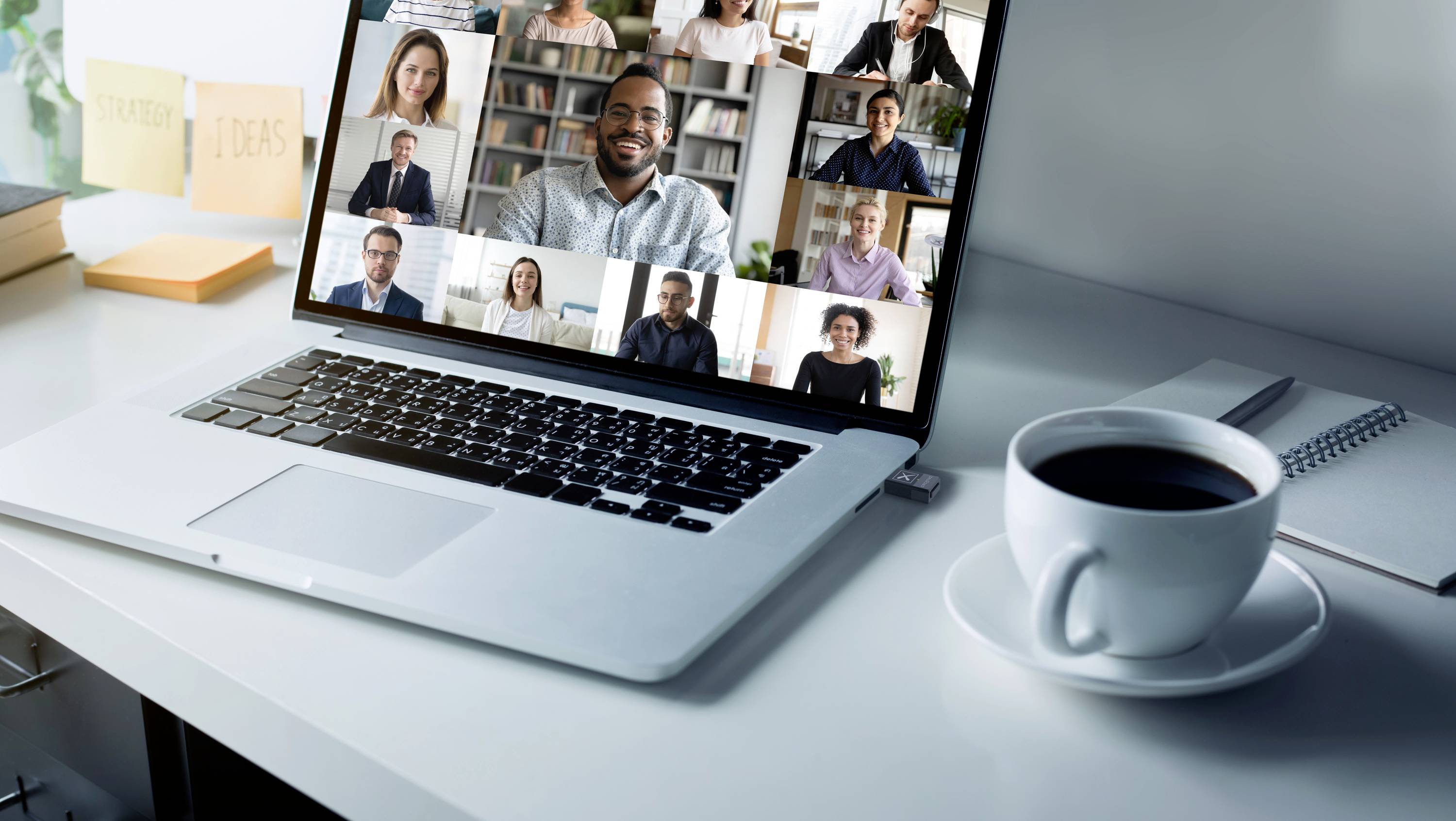 Ein Laptop zeigt eine Videokonferenz mit mehreren Personen auf dem Bildschirm. Daneben stehen Kaffee und Notizblock auf dem Tisch.