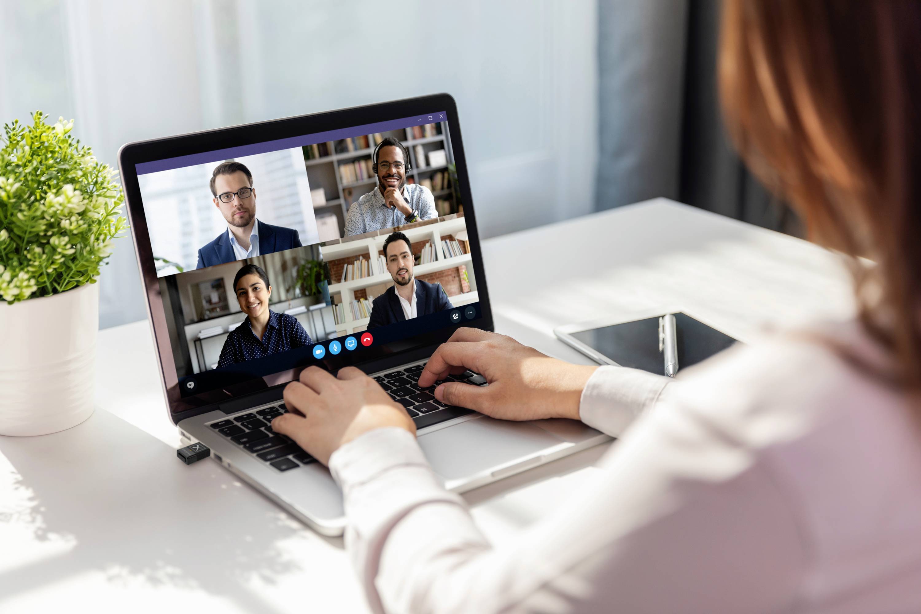 Person nimmt an einem Online-Meeting mit vier Teilnehmern über einen Laptop teil, auf einem Schreibtisch mit einer Pflanze und einem Notizbuch.