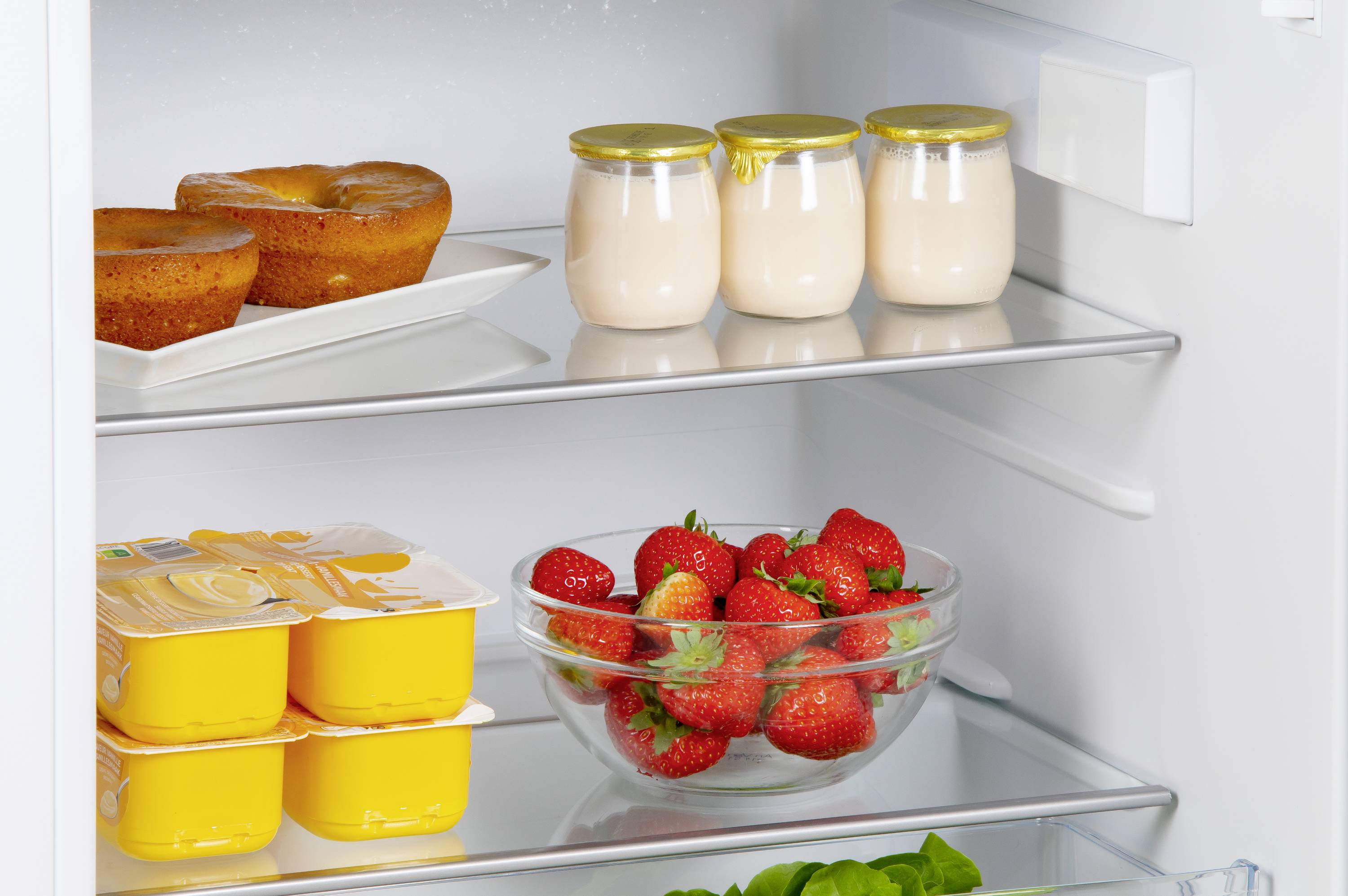 Offener Kühlschrank zeigt zwei Kuchen, drei Joghurts im Glas, Puddingbecher, eine Schüssel mit Erdbeeren und frischen Spinat.