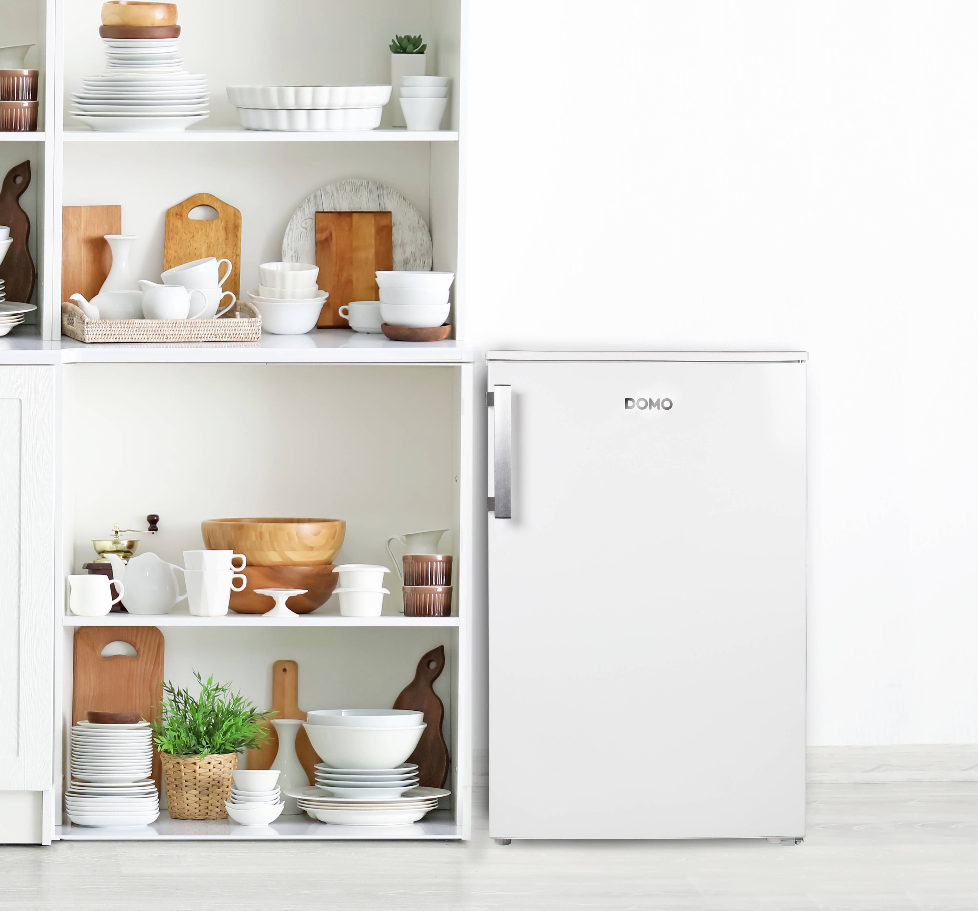 Ein weißer Kühlschrank rechts neben einem offenen Regal mit Geschirr, Töpfen und Pflanzen. Minimalistisches Küchendesign.