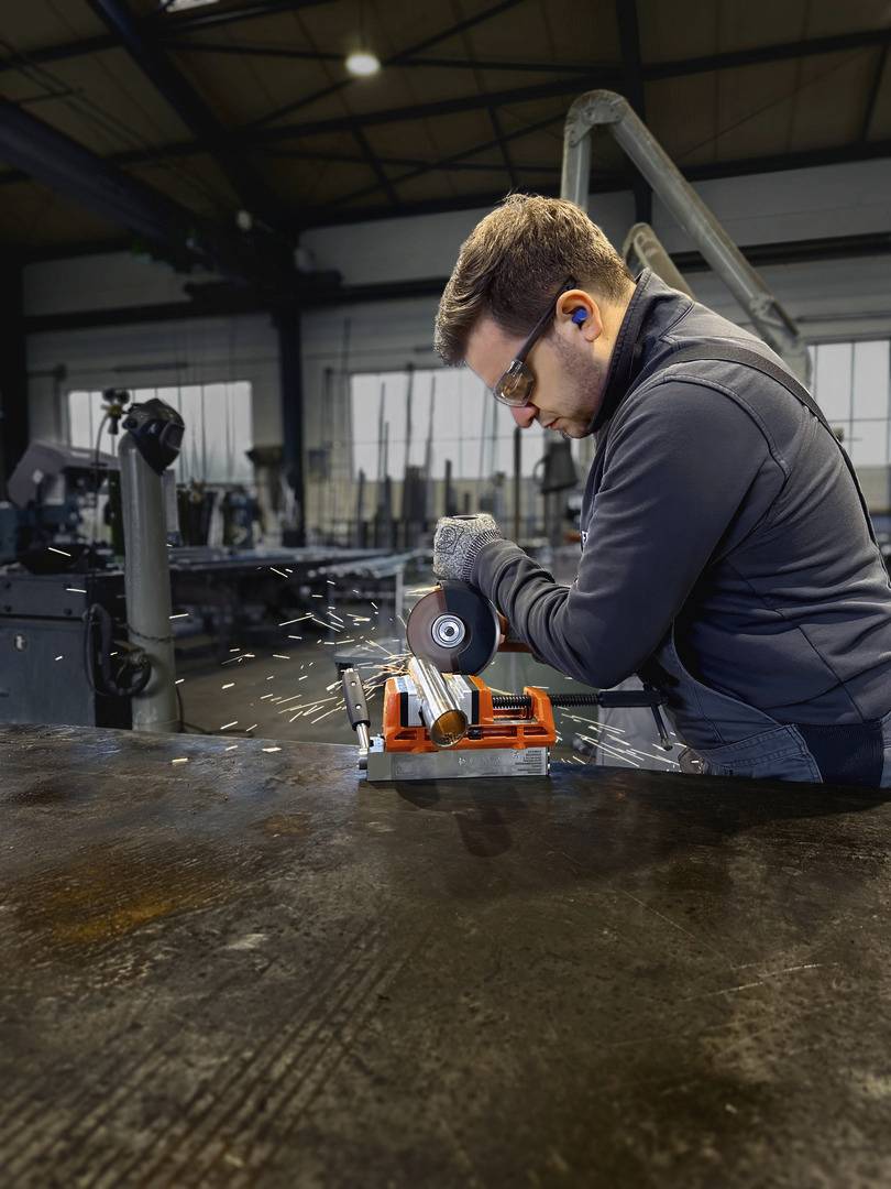 Ein Mann mit Schutzbrille und Gehörschutz schneidet Metall mit einer Schleifmaschine in einer Werkstatt. Funken fliegen.