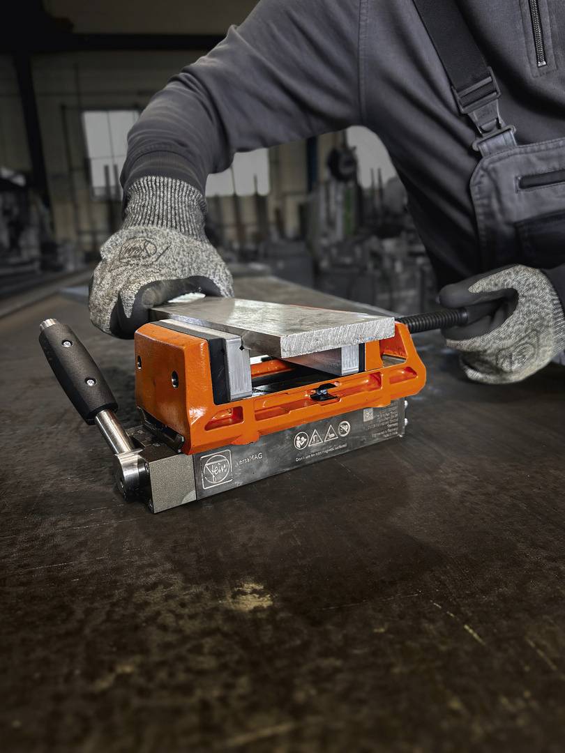 Eine Person arbeitet in einer Werkstatt mit einem magnetischen Hebezeug, das ein großes Metallstück anhebt.