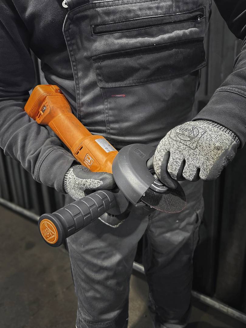 Eine Person in grauer Arbeitskleidung hält einen orangefarbenen Winkelschleifer in beiden Händen, bereit zum Einsatz.