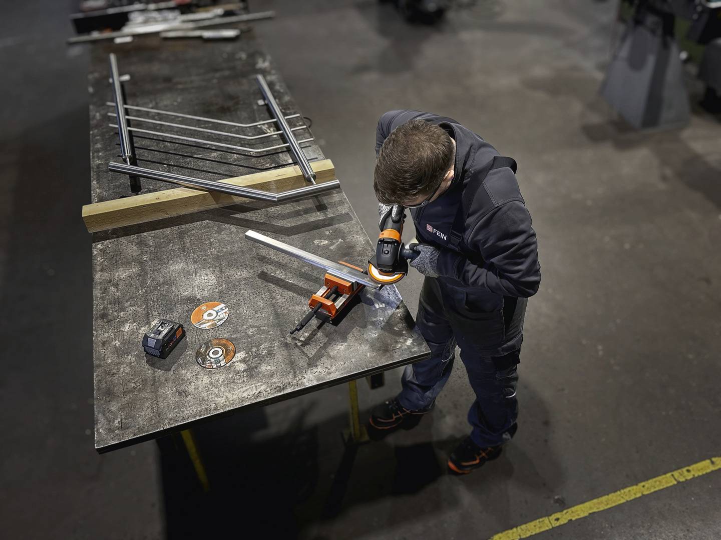 Eine Person in Arbeitskleidung bearbeitet ein Metallstück mit einer Bohrmaschine auf einem Werkbank. Holz- und Metallstücke sind um ihn herum platziert.