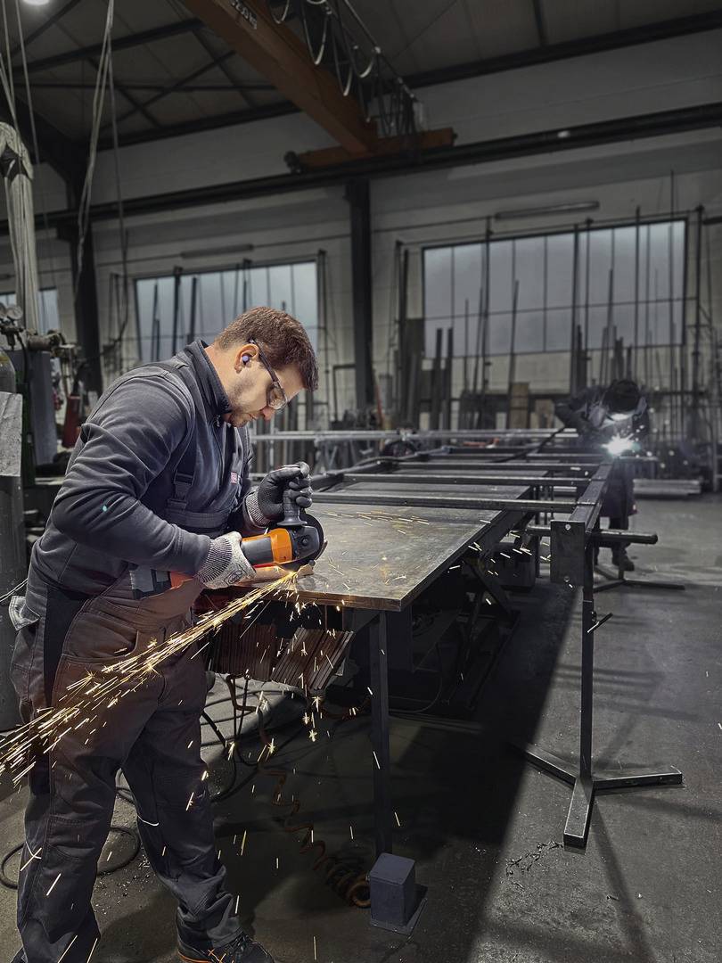 Ein Mann in Arbeitskleidung schleift Metall in einer Werkstatt. Funken fliegen von der Schleifmaschine. Im Hintergrund ist schweres Gerät.