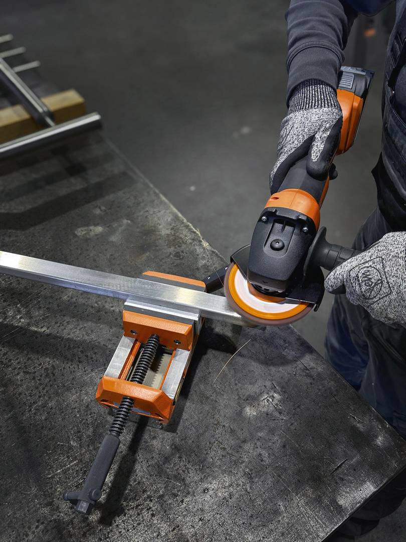 Person schleift mit einer Winkelschleifer-Maschine Metallkanten, Metallstück in Schraubstock fixiert, trägt Arbeitshandschuhe.